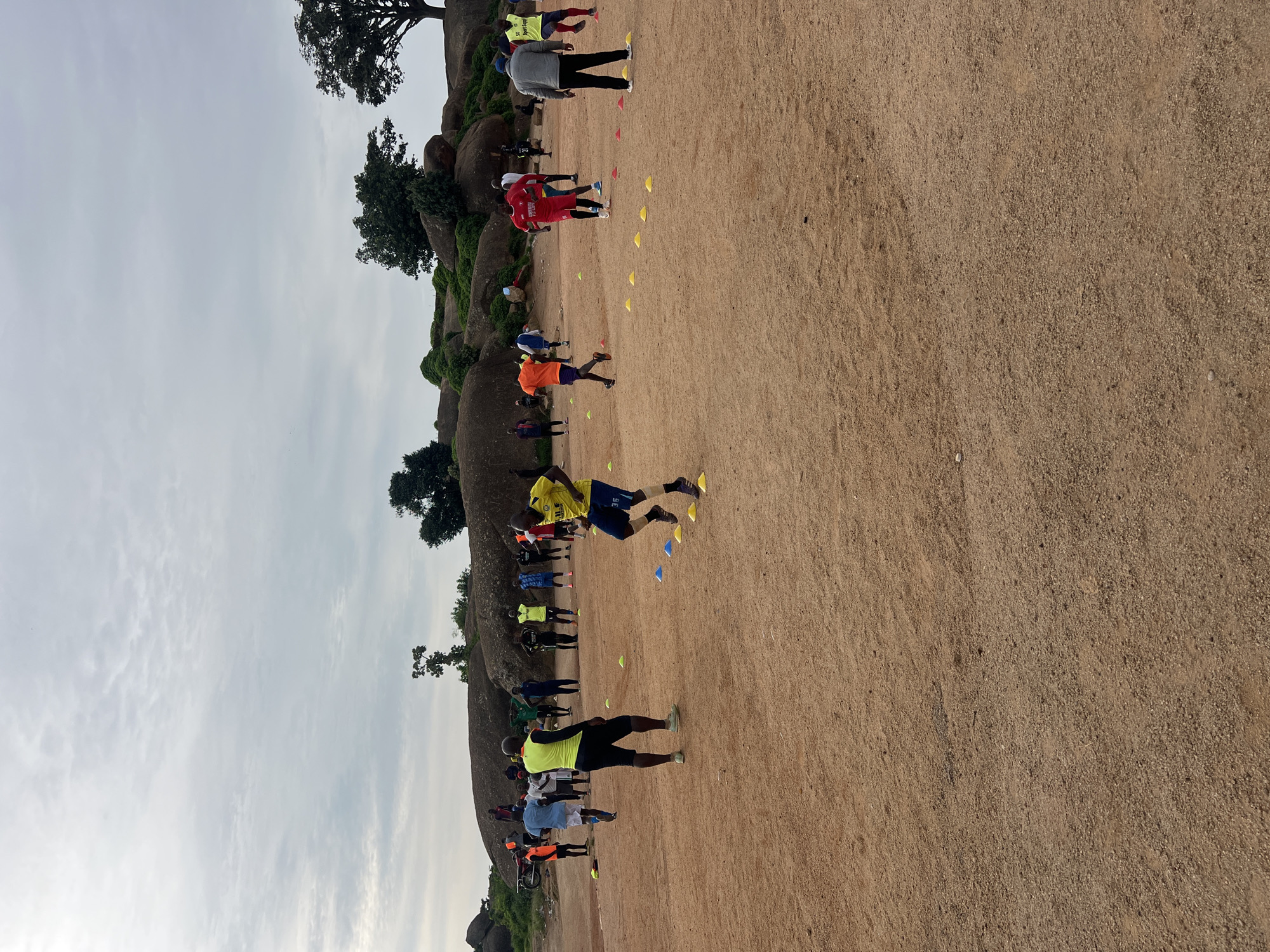 The image captures an outdoor scene in Jos, Nigeria, likely a training ground. A group of men, dressed in athletic wear, are engaged in what appears to be a football or agility training session. They are spread across a dusty, reddish-brown surface, with some individuals actively running and others standing. Several brightly colored cones, yellow and blue, are strategically placed on the ground, suggesting a drill or obstacle course.
In the background, large rock formations, characteristic of the Jos Plateau, dominate the landscape. These rocks are partially covered with green vegetation, and scattered trees are visible, including some larger ones towards the right of the frame. The sky is overcast with a light gray cloud cover. A motorcycle is parked near the rock formations on the left side of the image. The overall impression is a functional training area set against a natural, rocky backdrop.