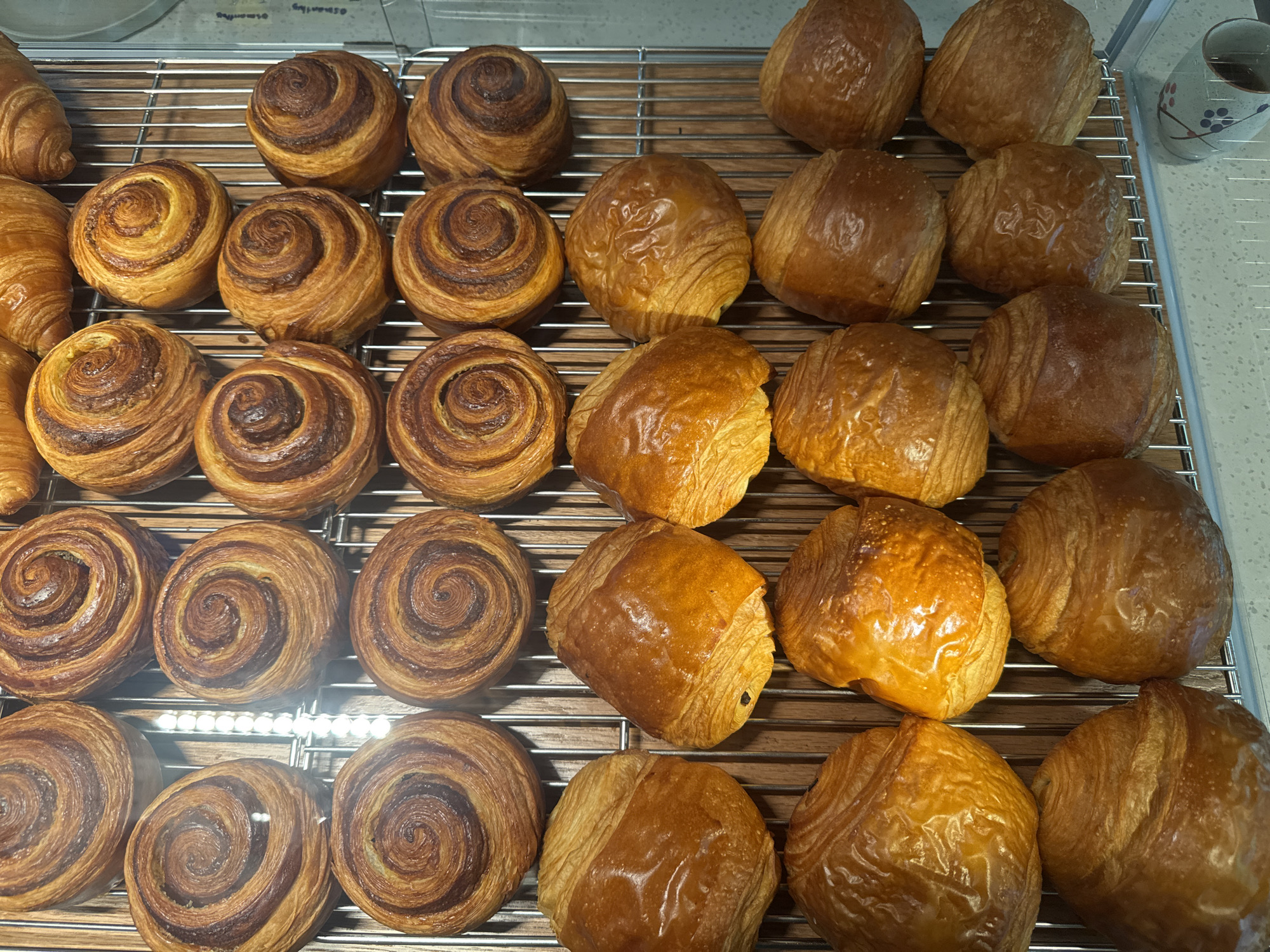 The image shows a close-up, overhead view of a rack of baked goods, likely pastries, arranged in rows. The items are golden brown and appear to be laminated, with visible layers. There are two distinct types of pastries: round, swirled cinnamon roll-like pastries and more rectangular, folded pastries that resemble croissants or pain au chocolat. The pastries are displayed on a silver wire rack over a wooden surface. The lighting is bright, casting a slight glare on the glossy surfaces of the pastries. There is a white reflective surface to the left of the pastries and a blurred background that appears to be a kitchen or bakery setting. There is no visible indication of the city of Hoi An, Vietnam, nor are there any people or actions depicted in the image. The scene is static and focuses solely on the arrangement of baked goods.