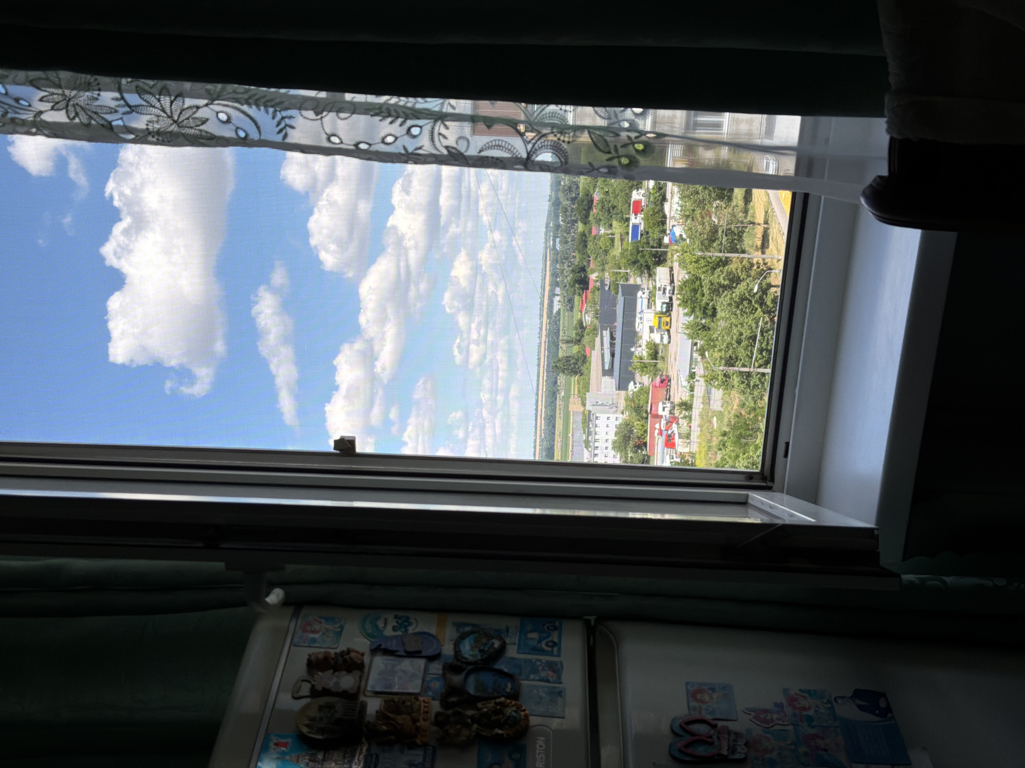 This image is a candid, outdoor shot taken from an interior perspective, looking out through an open window. The window frame is a prominent element, with a decorative sheer curtain visible at the top, featuring an embroidered floral pattern. Beyond the window, a bright, sunny day unfolds with a clear blue sky filled with fluffy white clouds.
The scene outside depicts a suburban or rural landscape. There are several buildings with red and white roofs, interspersed with lush green trees. A road or pathway is visible, with a few vehicles, including what appear to be trucks, parked or driving. The overall setting suggests a residential area with some commercial or industrial elements. The lighting indicates it is daytime, likely late morning or early afternoon, given the strong sunlight and clear skies.
The interior foreground of the image shows part of a refrigerator, adorned with various magnets and possibly some decorative decals. These magnets appear to be small trinkets, some resembling miniature buildings or landmarks, while others are more abstract or pictorial. One visible text on the refrigerator reads "ARISTON."
The image conveys a sense of a typical day, looking out from a home into the outside world. The weather is pleasant, characterized by sunshine and clouds. There are no people clearly visible, and the focus is on the scene outside and the items within the immediate interior. The perspective is slightly tilted, as if the photograph was taken from a casual angle.