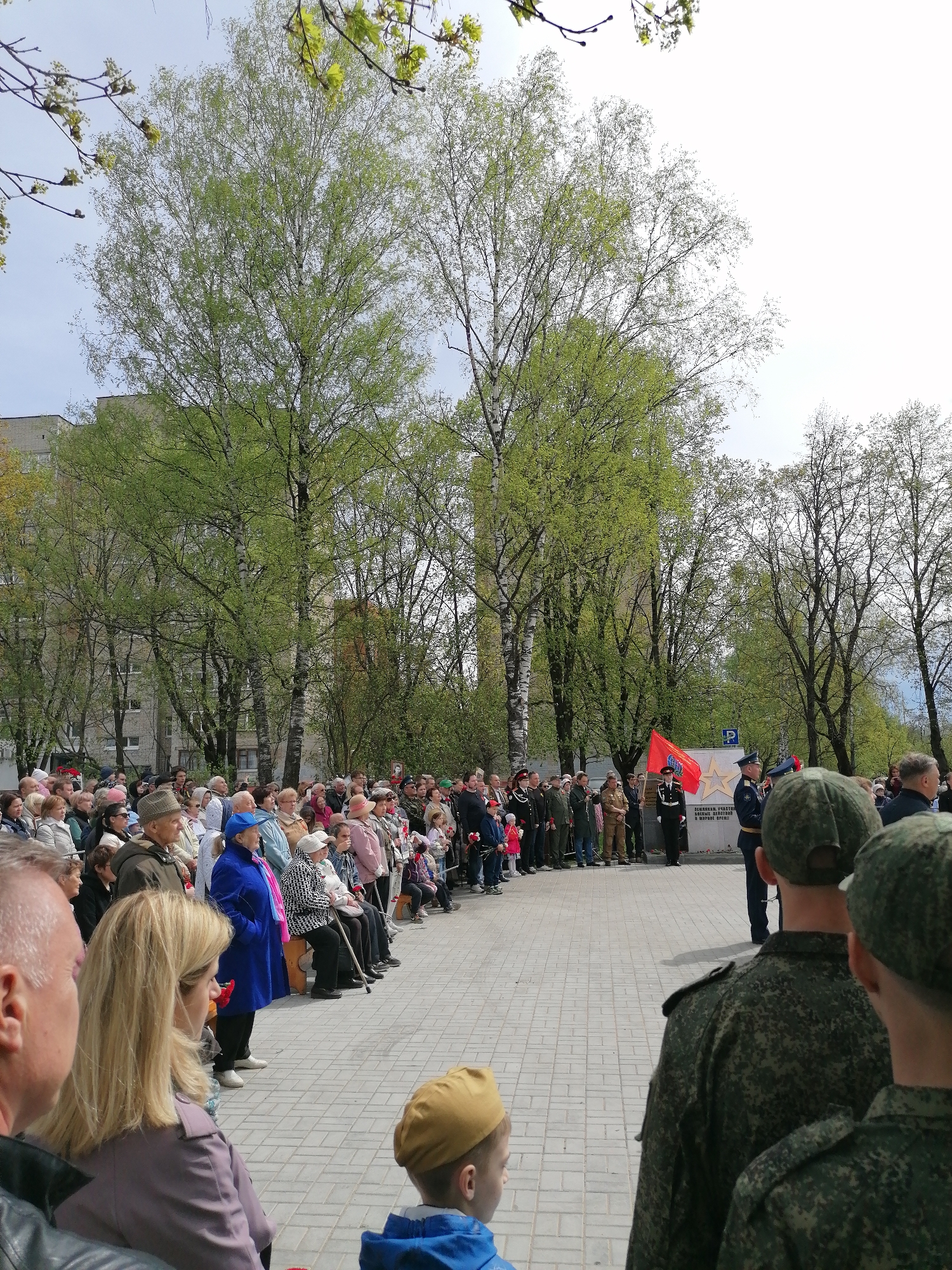 A crowd of people is gathered in a paved city square, likely for a ceremony or event. In the background, there are several mature trees with green foliage, and a multi-story building can be partially seen. A group of uniformed individuals are standing near what appears to be a monument or marker, and a red flag is visible. A small group of people in military uniforms are in the foreground, likely part of the honor guard or participating in the ceremony. The weather appears to be overcast with some hints of daylight breaking through the clouds.