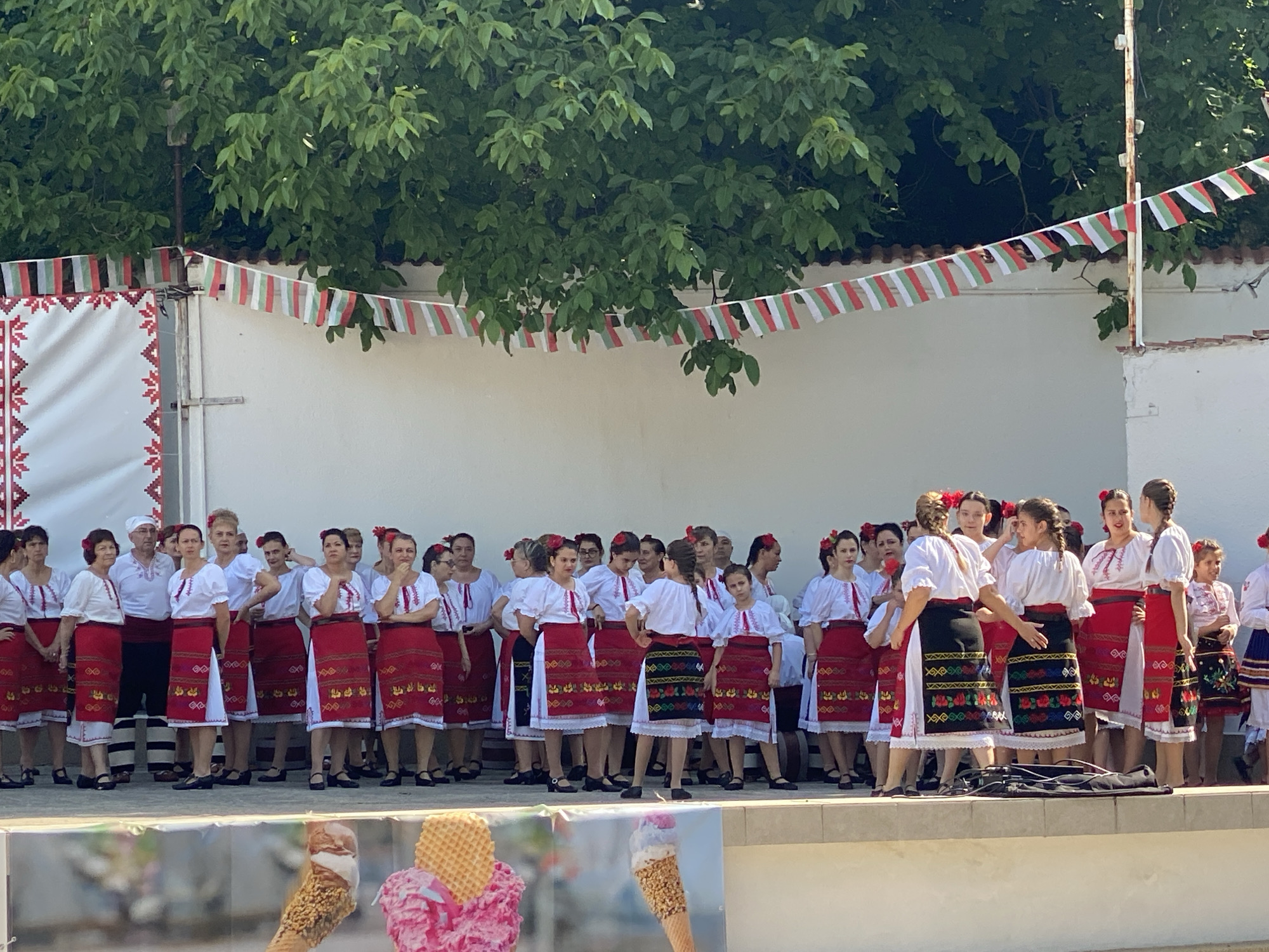 The image captures an outdoor cultural event in Pleven, Bulgaria, under bright daylight conditions. Approximately 30-35 individuals, predominantly women and girls with one adult male, are gathered on a raised, light-colored stage or platform. They are uniformly dressed in traditional Bulgarian folk costumes, which include white blouses with embroidered sleeves, red aprons or skirts featuring intricate geometric or floral embroidery (some aprons on younger girls are black with colorful floral patterns), and white underskirts. Many of the females have red flowers or ribbons adorning their hair. The male figure and some females wear red sashes around their waists.
The background consists of a plain white wall that serves as a backdrop for the performers. Above this wall, dense green foliage from trees is visible, indicating an outdoor setting. A string of small rectangular flags, displaying vertical white, green, and red stripes (the colors of the Bulgarian flag), is strung horizontally across the upper portion of the scene. On the far left, a white fabric panel or banner, decorated with a red geometric embroidered border, is affixed to a structure. In the bottom foreground, a partial banner displays photographic images of ice cream cones. A black, rectangular carrying case, possibly for a musical instrument, rests on the stage floor in the center-right. The individuals on stage are standing in various poses, suggesting they are either awaiting a performance, have just completed one, or are posing for an event.