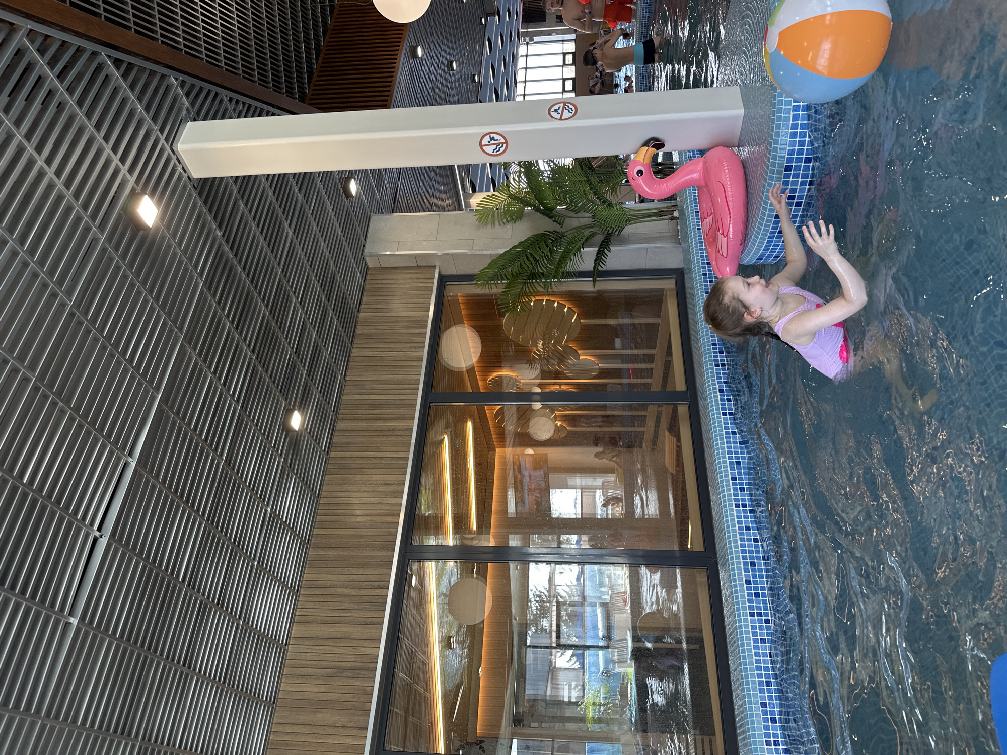The image shows a young girl swimming in a pool next to an inflatable pink flamingo and an orange and blue beach ball. The pool has a blue mosaic tile edge. A white structure with warning signs is positioned near the pool. A palm plant is situated between the structure and the window. The background features a large window with reflections, and a wall with a textured gray surface. The location appears to be an indoor water park, likely in Votkinsk, Russia. Other people are visible in the background, suggesting a public swimming area.