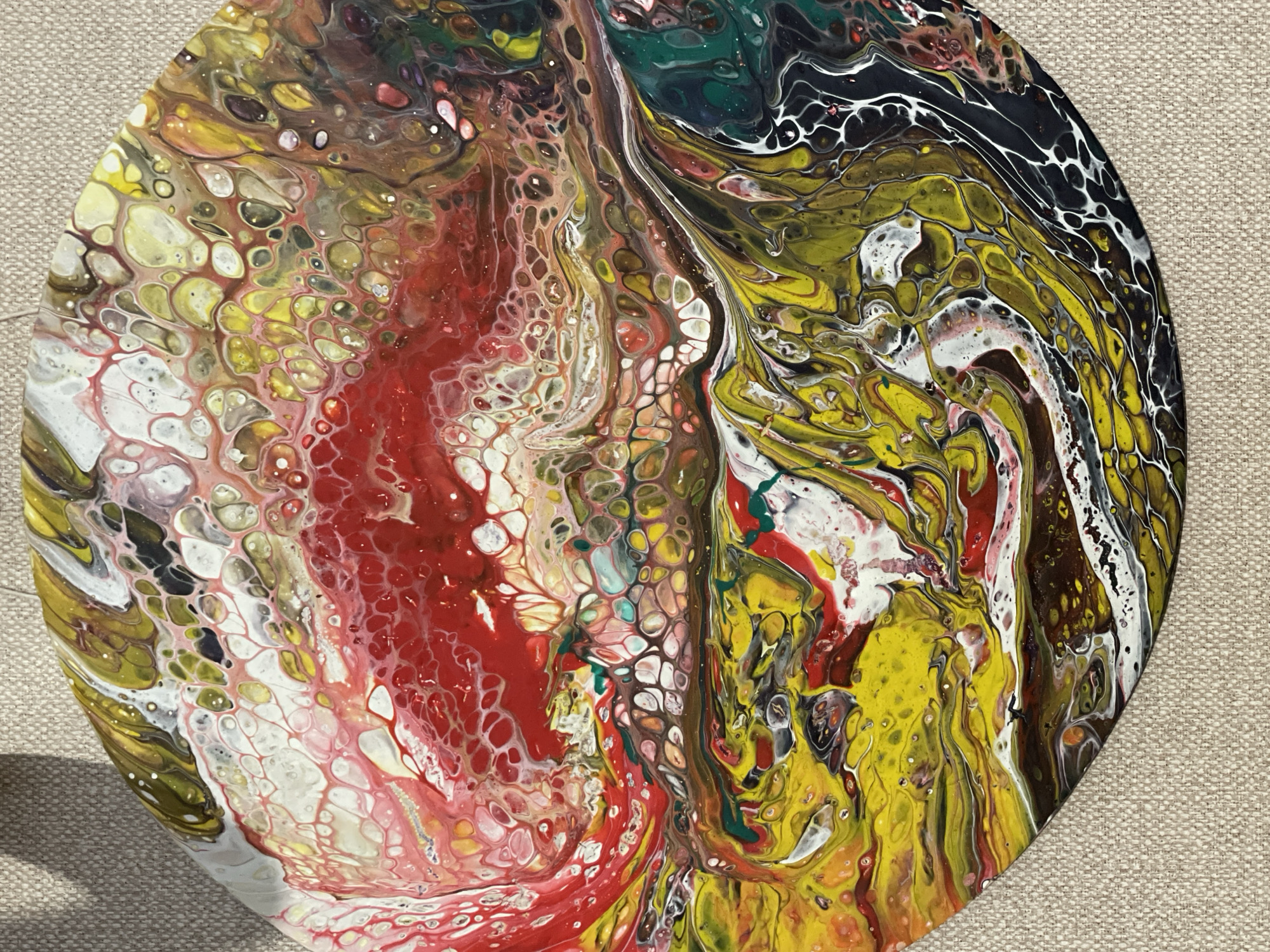 A circular abstract acrylic pour painting is presented on a textured surface. The painting features a dynamic mix of colors, primarily red, yellow, white, and black. The red forms a central mass with cellular patterns, while yellow dominates the right side with swirls and string-like formations. White accentuates the edges of the red and yellow areas. Black is concentrated in the upper right corner, interlaced with white lines to create a marbled effect. The painting is placed on a neutral-colored, possibly linen-textured background. A subtle shadow suggests a light source from the left. The painting appears to be a close-up shot, capturing the details of the acrylic pour technique.
The location of Zheleznogorsk, Russia is irrelevant to the visual details in this image.