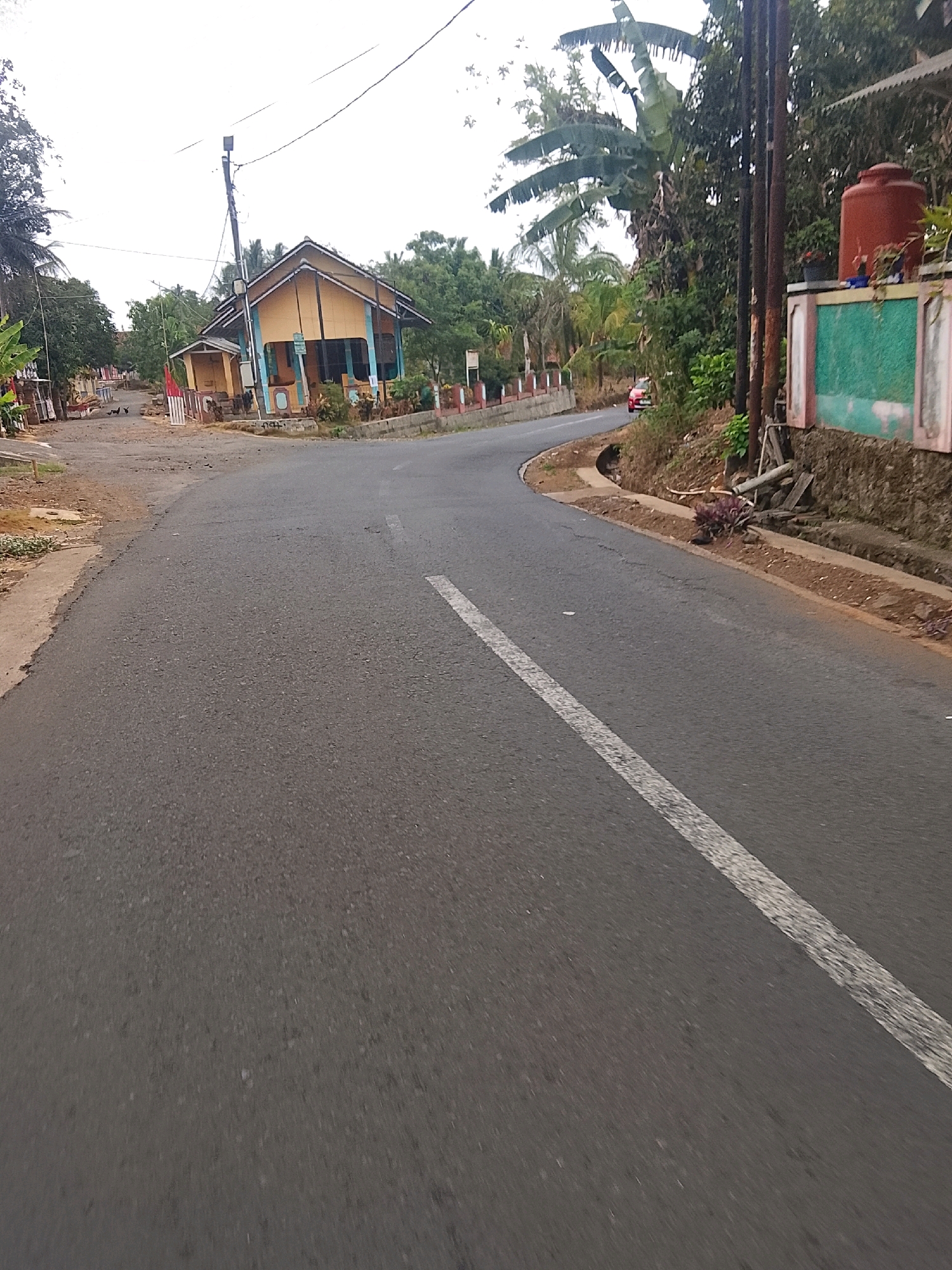 A paved road curves through a semi-rural area in Banjar, Indonesia. The road has a white dashed line down the center, indicating it is a two-way street. On the left side of the road, there is a small yellow and blue building with a porch. In front of the building, several chickens are foraging on the ground. Further down the road on the left, there are more buildings and trees. On the right side of the road, there is a retaining wall with a green and pink facade. A large orange water tank sits on top of the wall. Dense vegetation, including palm trees, lines the right side of the road. A red car is visible in the distance, driving on the road. The sky is overcast, suggesting cloudy weather.