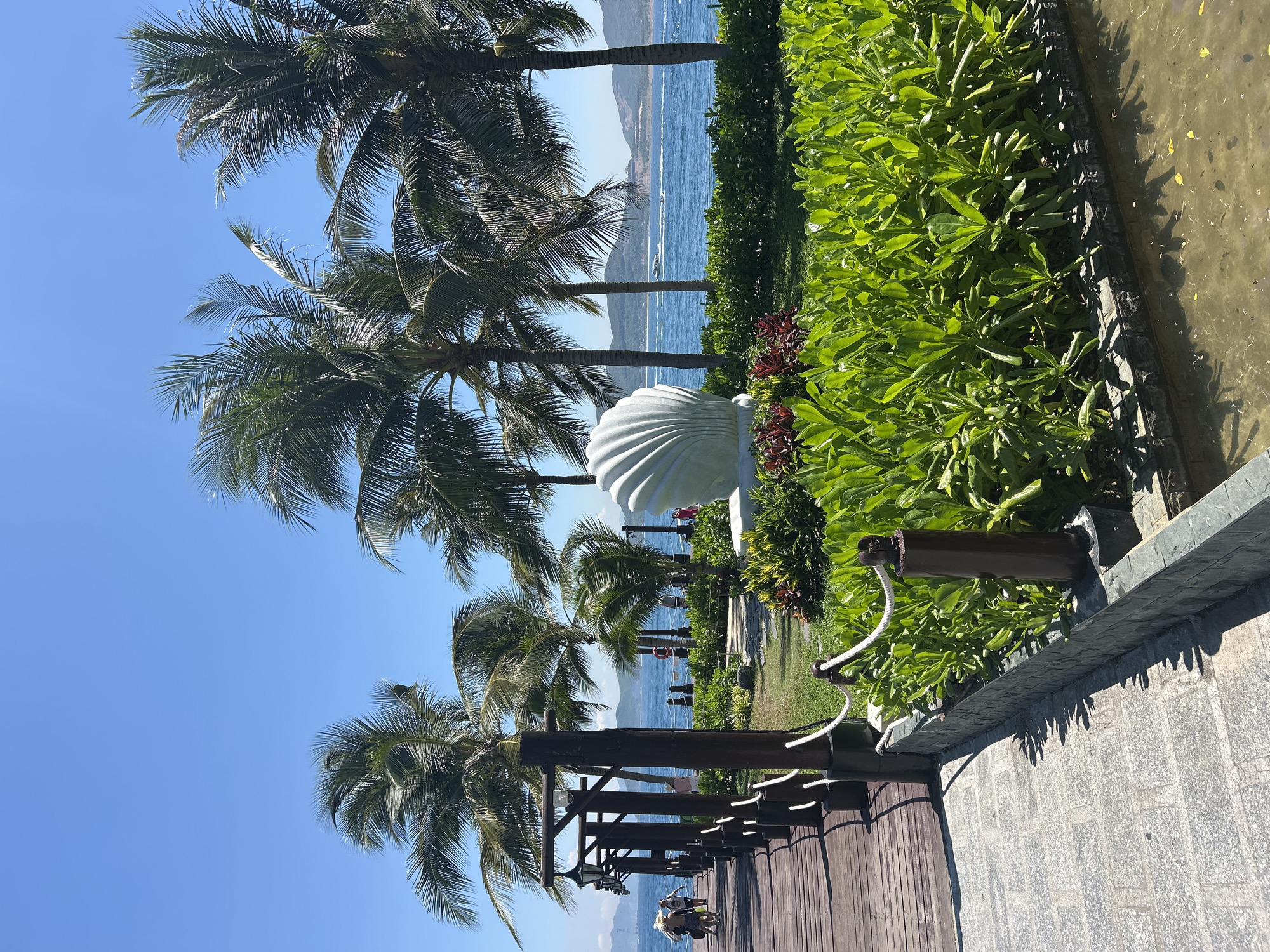 The image depicts a pier extending into the ocean in Nam Nha Trang Ward, Vietnam. The pier is constructed of weathered wooden planks and lined with dark wooden posts supporting lamps and white ropes. To the right of the pier, there is a meticulously manicured garden featuring lush green bushes and red-leafed plants. A large white sculpture resembling a seashell is prominently displayed within this garden area. Palm trees are scattered throughout the landscape, their fronds swaying gently. The ocean is a vibrant blue, with a few small boats visible in the distance. The sky is clear and a bright azure. In the background, hills and mountains form the coastline. Several people are visible on the pier, walking towards the end, and some are seen in the distance near the garden area. The overall scene suggests a resort or recreational area by the sea.