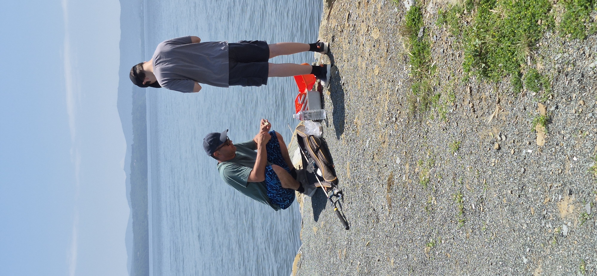 Two individuals are by the water on a rocky shore. The person on the left is a man wearing a green t-shirt, blue patterned shorts, a dark baseball cap, and sunglasses. He is crouched down and appears to be preparing fishing bait. To his left is a fishing rod, a fishing bag, and a plastic bag. To his right is a bottle of water and a red container. The person on the right is a male wearing a grey t-shirt, dark shorts, and a black baseball cap. He is standing and looking down, possibly at his phone or something in his hands. In the background, there is a large body of water with distant, hazy hills. The sky is clear and blue. This scene does not appear to be in Vladivostok, Russia, as the landscape and attire of the individuals suggest a different, likely warmer, location.
