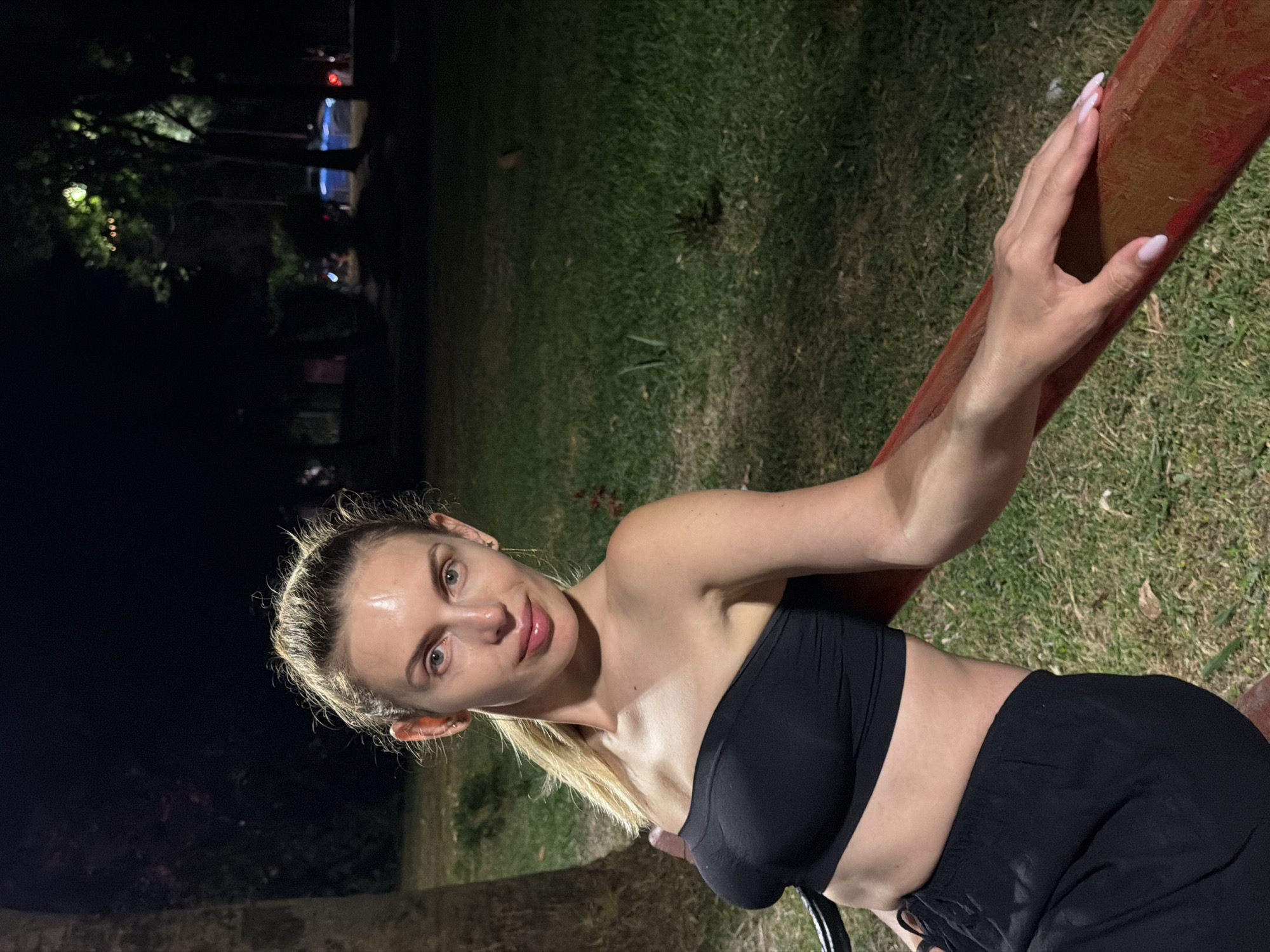 A woman with blonde hair pulled back is shown from the chest up, leaning on a red railing. She is wearing a black bandeau top and black pants. Her head is slightly tilted, and she is looking at the camera with a slight smile. The background is dark, with some blurred greenery and lights in the distance. The image appears to have been taken at night, possibly in a park or outdoor recreational area. The woman has on light pink nail polish. The scene is illuminated by artificial lights, creating some shadows and highlights on her skin and hair.