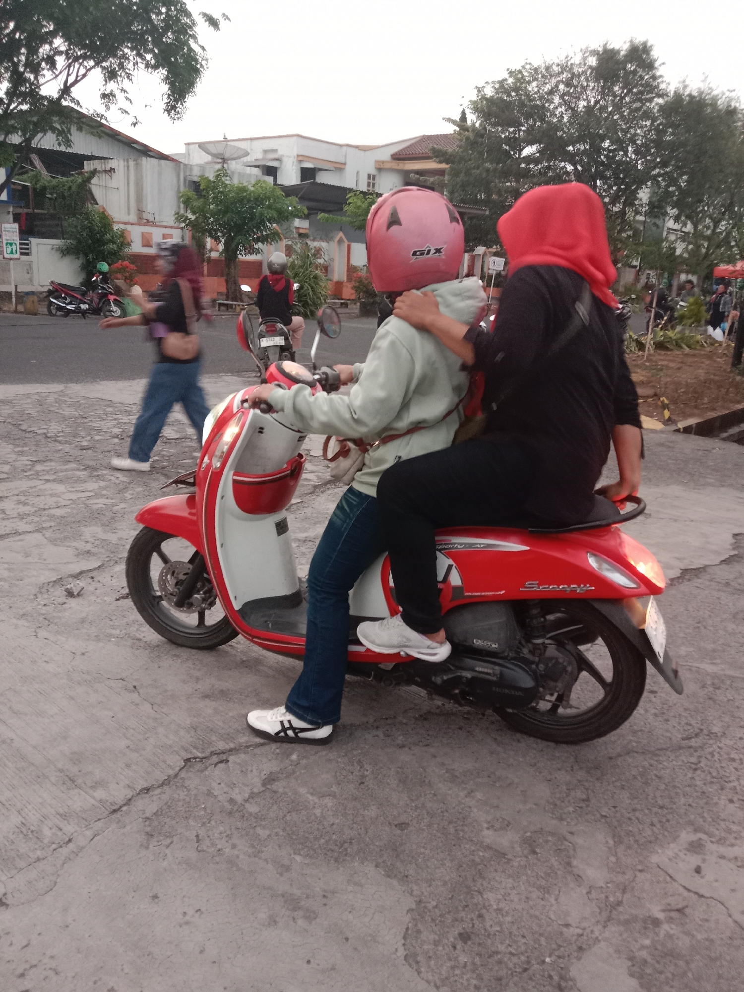 In Semarang, Indonesia, two individuals are seen riding a red and white scooter on a paved street. The person in front is wearing a light green jacket and a pink helmet with the brand "GIX" visible. The person in the back is wearing a black top and a red headscarf. Both are wearing blue jeans. The scooter has the word "Scoopy" written on the side. In the background, there are buildings with a satellite dish on one of them, trees, and other motorcycles parked along the street. The lighting suggests it is either early morning or late afternoon.