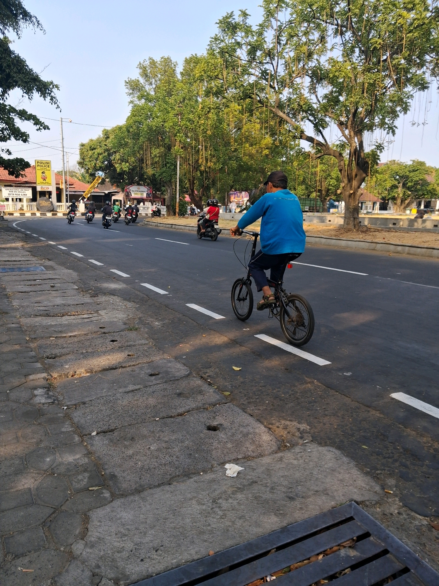 A man is riding a bicycle on the right side of a road in Surakarta, Indonesia. The man is wearing a blue long-sleeved shirt and dark pants. He is riding a small, black bicycle with thin tires. The road is asphalt with white lane markings. There are several motorcycles in front of him, also riding on the road. On the left side of the road, there is a sidewalk with a grate. Behind the sidewalk, there is a police station with signs in Indonesian. The area is lined with trees, some of which have long, hanging vines. The sky is clear and blue.