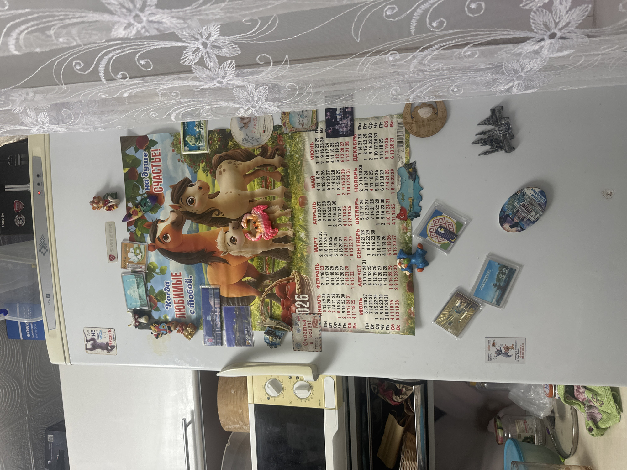 The image shows the exterior of a white refrigerator, likely in a kitchen. The refrigerator door is covered with various magnets and a calendar. A large calendar featuring cartoon horses with the text "Когда любимые с тобой" (When loved ones are with you) and "На душе счастье!" (Happiness in your soul!) is prominently displayed. Below this, a monthly calendar grid for an unspecified year is visible, showing dates for several months. There are also numerous smaller magnets, some depicting landmarks like the Taj Mahal and Sagrada Familia, and others with cartoon characters and text. A small figurine of a blue dog is also attached to the refrigerator. To the left of the refrigerator, a microwave is partially visible. The background shows a wall with a textured pattern and a white curtain with floral embroidery. The scene appears to be an indoor domestic setting, with no specific indication of the city of Загородный, Russia, beyond the possibility that the refrigerator is located there.