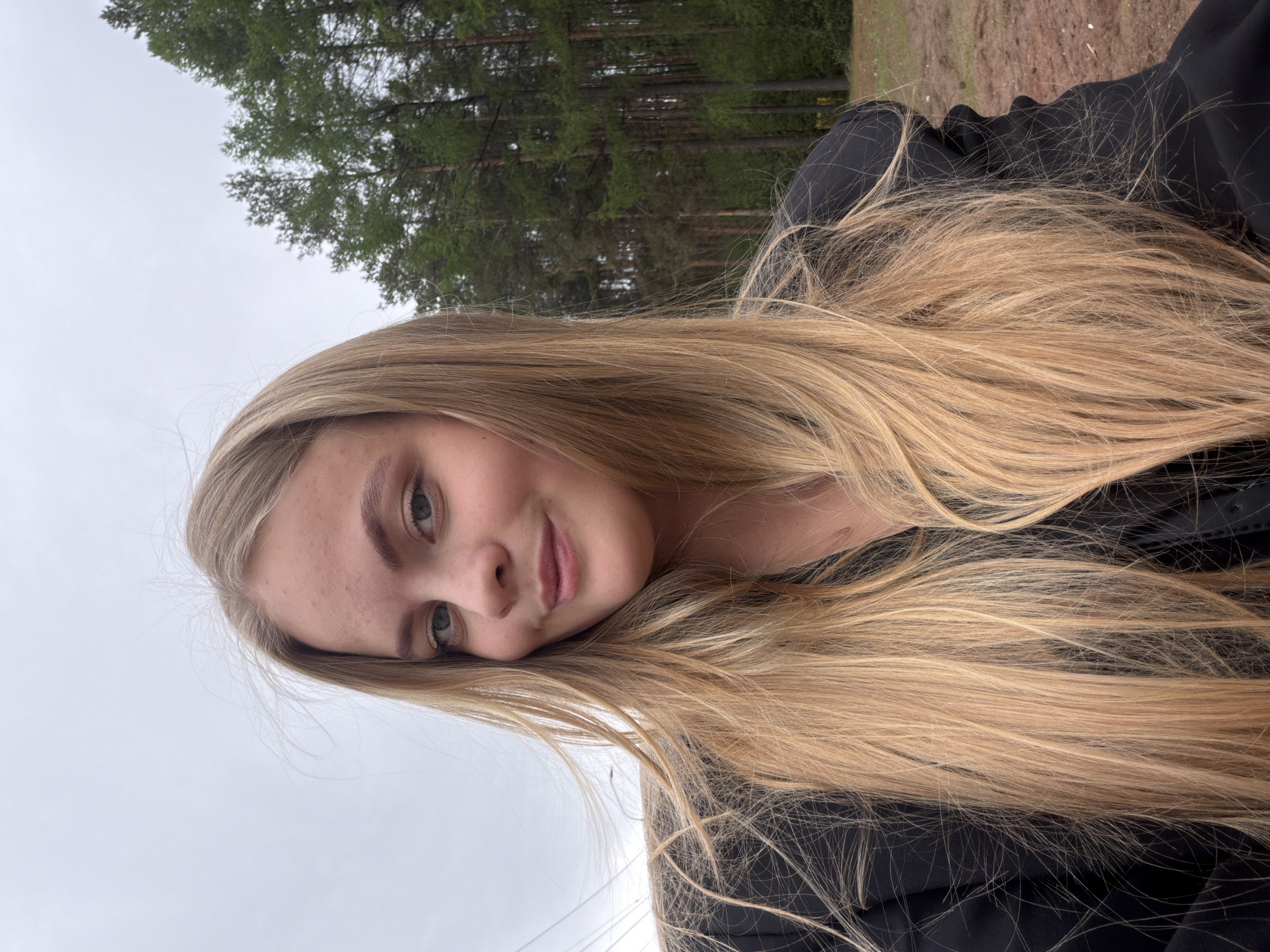A young woman with long blonde hair is posing outdoors. She has fair skin, light eyes, and is wearing a black top. The background includes trees and a gray sky. The specific location within Izhevsk, Russia, cannot be determined from the image alone.