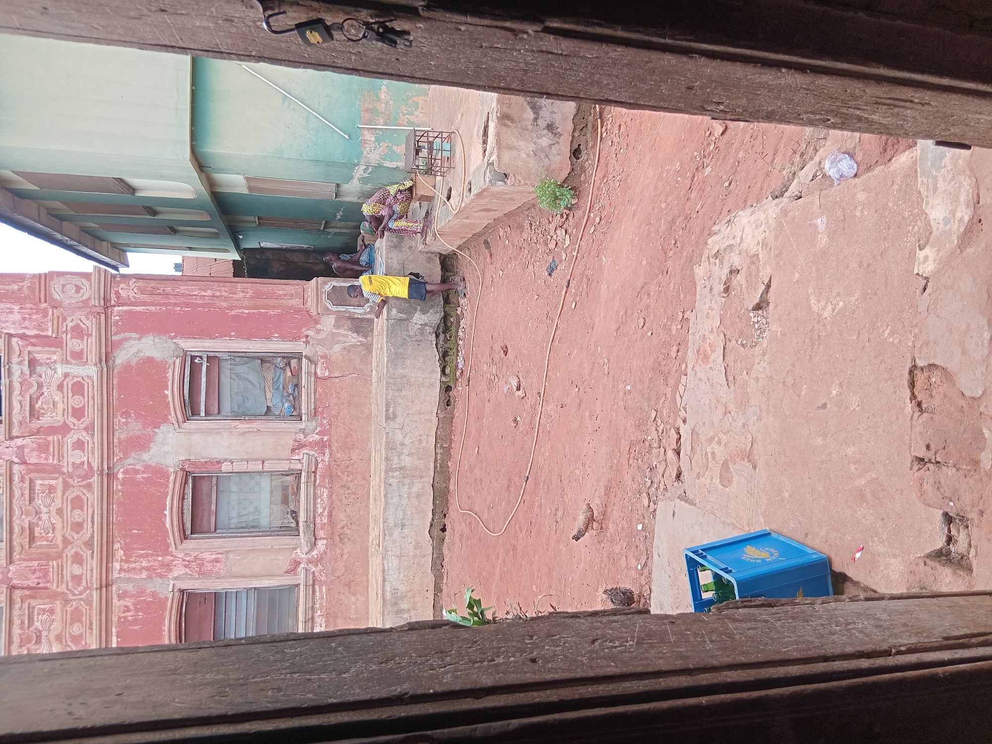 From an elevated, partially obstructed view, the image captures a courtyard scene in Shagamu, Nigeria. The architecture includes a red-brick building with ornate detailing around the windows, and a building painted with light green and turquoise accents is also visible. A group of people are gathered near a concrete ledge, including one shirtless individual. There is an orange hose snaking across the red-earth ground. A blue plastic crate is positioned near the bottom of the frame. Two chickens are visible, one near the hose and another on the ground. The view is framed by the edges of what appears to be a wooden window or doorway.