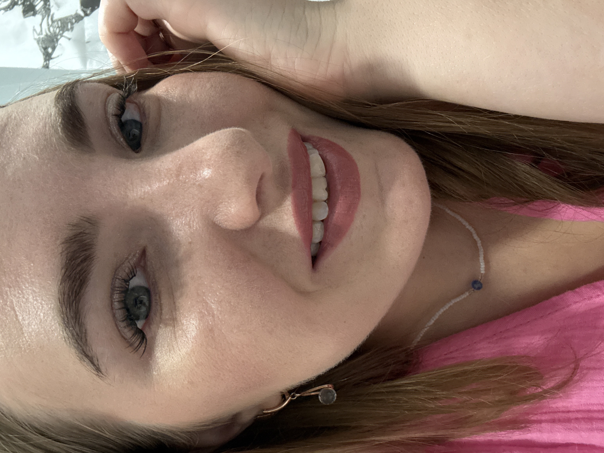 The image is a close-up, selfie-style portrait of a young Caucasian woman with light brown hair, blue eyes, and fair skin. She is wearing a bright pink top and a delicate beaded necklace with a blue pendant. She has on makeup, including mascara and lipstick. A small, dangling earring with a gray stone is visible on her left ear. Her right hand is near her face, with fingers touching her cheek. The background is blurred and appears to be indoors, possibly a room with a patterned curtain or wallpaper on the right side. There is no indication of the location being in Kazan, Russia.