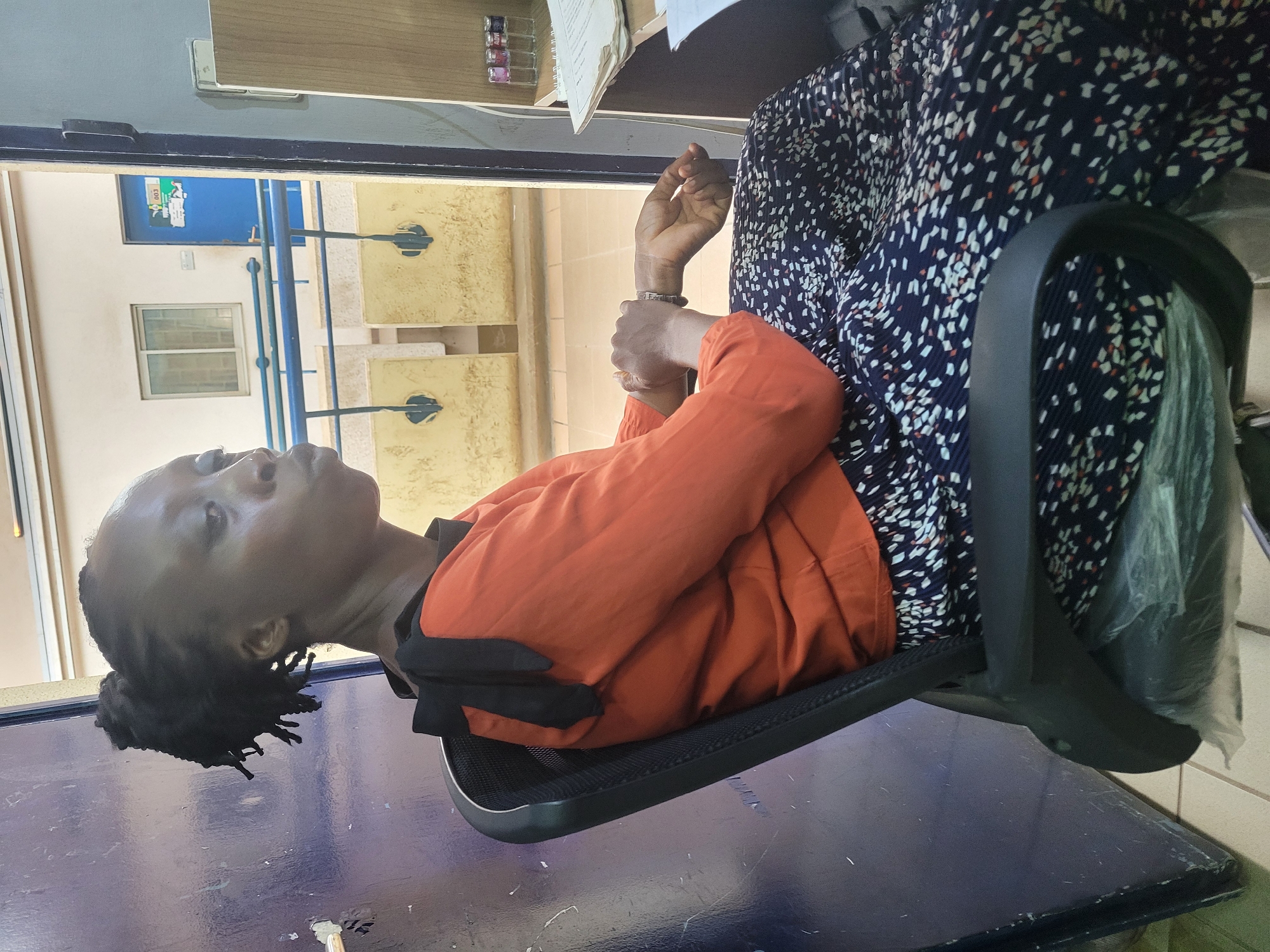 The image, rotated, depicts an indoor scene in what appears to be an office or institutional setting in Abuja, Nigeria. An adult female with dark skin and dark dreadlocks is the primary subject, seated in a black office chair with a mesh back. She wears an orange long-sleeved top and a dark blue skirt adorned with white and orange geometric patterns. She is looking upwards and slightly to her left, with her hands clasped together, suggesting a moment of rest, thought, or observation.
In the foreground, a dark blue table or desk surface is prominent. The background features light-colored walls and a multi-paned rectangular window, offering a glimpse of the exterior. Outside, blue metal rails or bars are visible, along with parts of another building structure. A blue door with a white sign displaying "003" is seen in the distance, indicating a room number. On a shelf or surface in the upper right, several small glass containers holding colored liquids are present; one container faintly shows the letters "ADW." Loose papers are also stacked nearby. The lighting suggests it is daytime. The setting implies a functional and possibly academic or administrative environment.