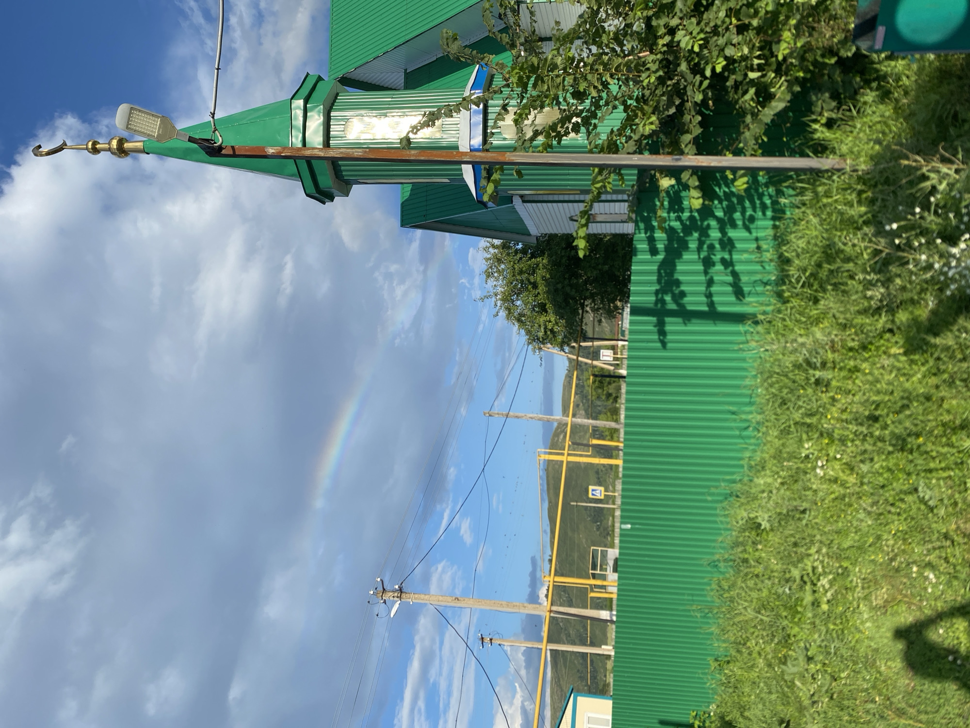 The image depicts a scene in Maloarslangulovo, Russia, featuring a mosque with a green roof and minaret topped with a golden crescent. A rainbow is visible in the cloudy sky. In the foreground, there is a bright green corrugated metal fence and lush green vegetation. Power lines and utility poles are present, along with a yellow pipeline. The overall environment suggests a rural or semi-rural setting.