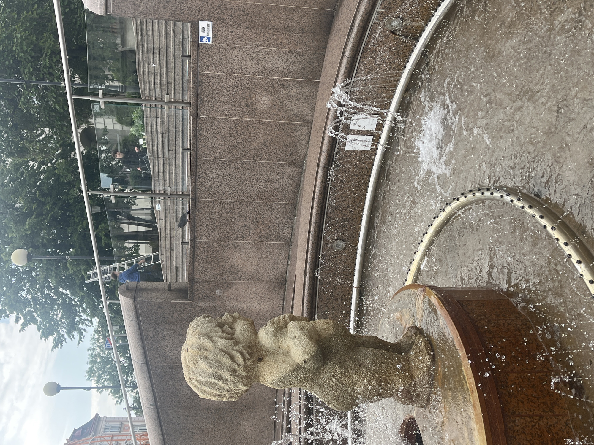 In a city fountain in Unknown, Unknown, a stone statue of a cherubic child stands with water spraying around it. The statue is positioned on a circular wooden and stone base. The fountain's basin is filled with splashing water. In the background, a textured granite wall rises, with a metal railing and glass panels above. Further back, there are steps leading up to a building, with trees and streetlights visible. Two individuals are present in the background; one is on a ladder near the steps, and another is seated on the steps. A pigeon is visible on the ledge in front of the granite wall, and another is on the steps. A sign indicating "OBIEKT MONITOROWANY" (Object Monitored) is affixed to the granite wall.