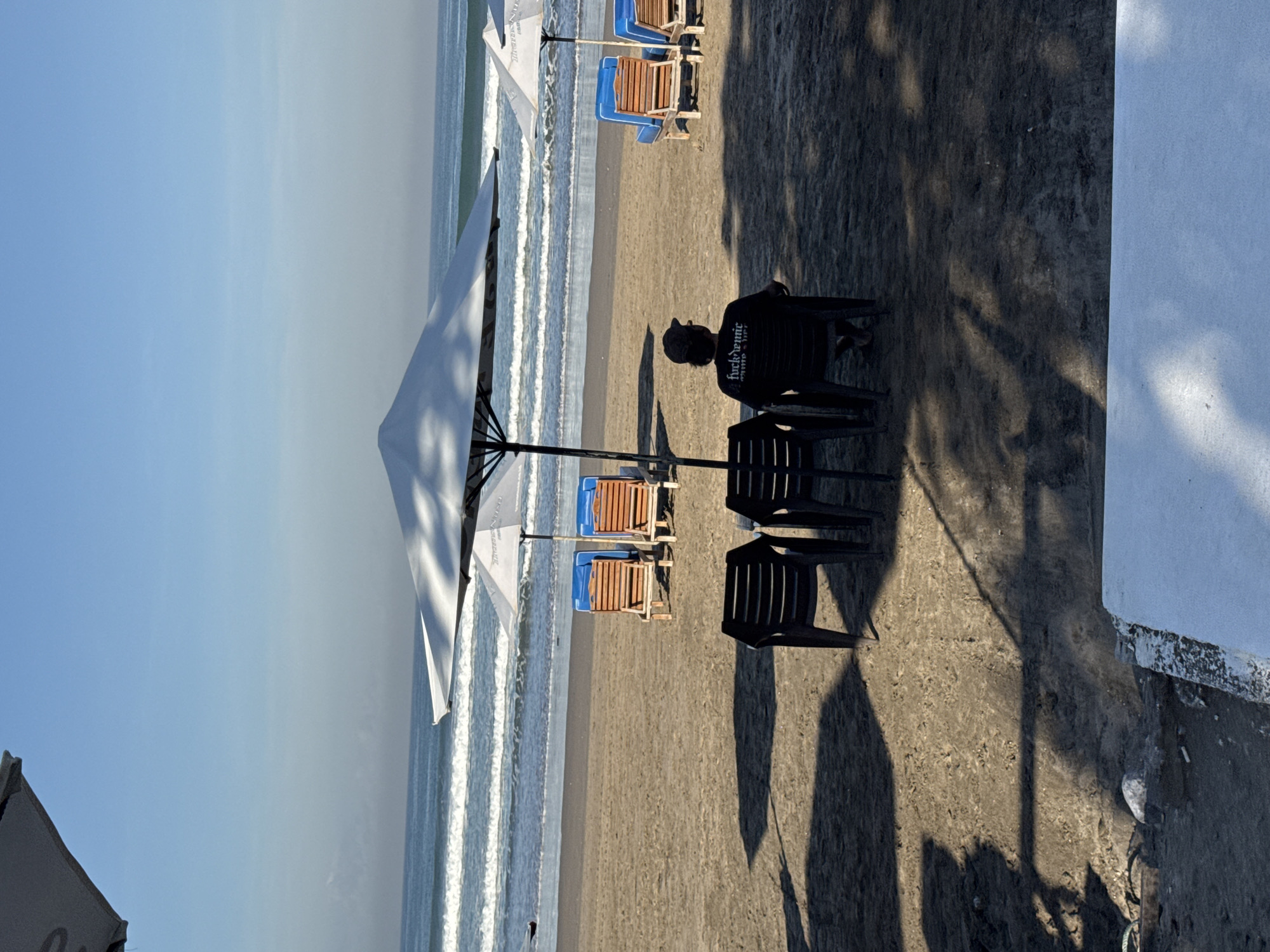 The image captures a beach scene in Kuta, Indonesia. The foreground contains beach chairs and an individual seated with their back facing the viewer. A large white beach umbrella provides shade. The background features the ocean with gentle waves, a sandy beach, and clear blue sky. A building is partially visible on the right side of the image.