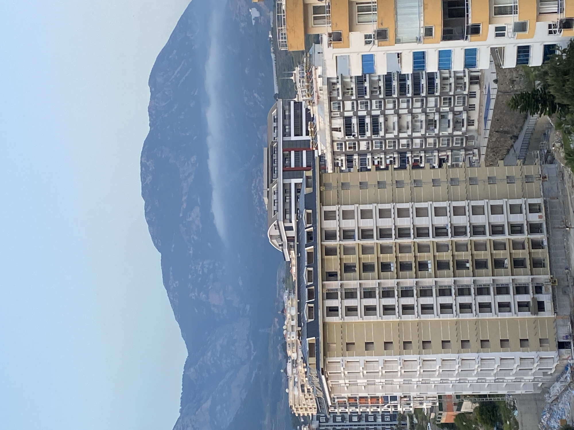 The image displays an urban landscape juxtaposed against a large mountain range, indicative of a coastal city like Alanya, Turkey. Multi-story buildings, likely a mix of residential apartments and hotels, dominate the right side of the frame, filling the foreground and midground. These structures exhibit repetitive patterns of windows and balconies, with facades in varying shades of beige, white, and some darker tones. Several buildings feature prominent blue-tinted windows. Elements suggesting ongoing construction or recent development, such as building materials and unfinished sections, are visible at the base and between some structures.
To the left and in the background, a vast mountain range rises dramatically. The upper parts of the mountains are partially enveloped by a soft, light mist or low clouds, creating a hazy effect. The sky above is a clear, light blue, suggesting a daytime scene, possibly early morning or late afternoon given the diffused illumination. Small clusters of green foliage, including some pine-like trees, are scattered amongst the buildings and on the lower mountain slopes. A small swimming pool can be faintly observed among the complexes. No people are visible, and there is no discernible text within the image. The scene captures a blend of dense human infrastructure and natural mountainous topography.
