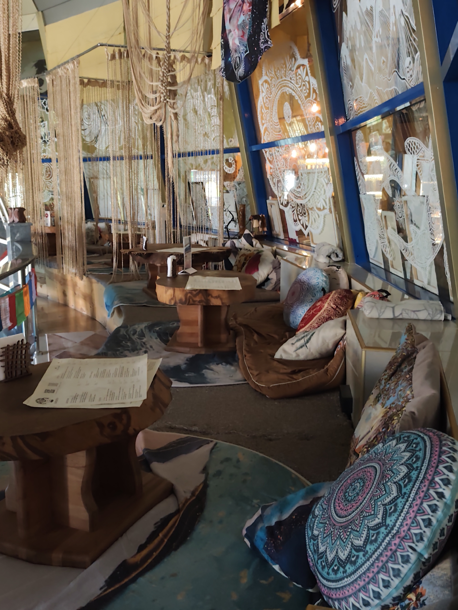 The image depicts the interior of a cafe, likely located in Sochi, Russia, characterized by a bohemian and artistic ambiance. The space is adorned with numerous macrame hangings and intricate white wall decals resembling mandalas and natural motifs, creating a visually rich and textured environment. Natural wood furniture, including tables and seating areas, is prominent throughout the scene. Cushions with vibrant, patterned covers, including one with a mandala design, are scattered around low seating areas, suggesting a relaxed and comfortable atmosphere. In the foreground, a wooden table holds a menu, and the floor is covered with rugs featuring abstract and wave-like patterns. The lighting appears to be warm and ambient, with visible light fixtures reflecting in the windows. The overall impression is one of a unique, cozy, and artfully decorated establishment.
