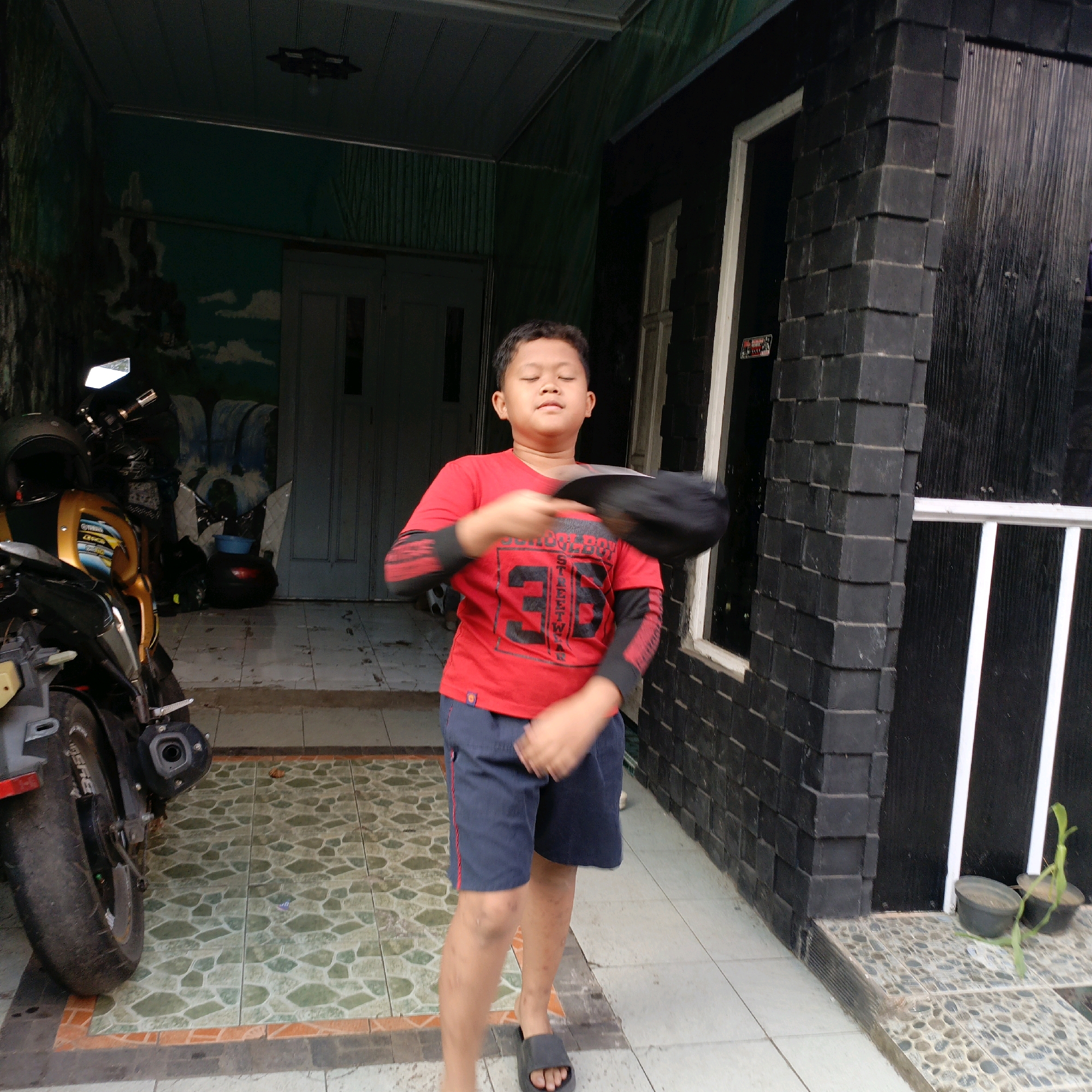 A young boy wearing a red t-shirt and dark blue shorts stands in front of a house with a black brick facade in Salatiga, Indonesia. He is in the process of throwing a black hat upwards with his right hand. His eyes are closed, and his mouth is slightly open. A motorcycle is parked to the left of the boy, partially obscuring a mural on the wall behind him. The ground is tiled with a mosaic pattern. The scene appears to be an outdoor entryway or porch.
