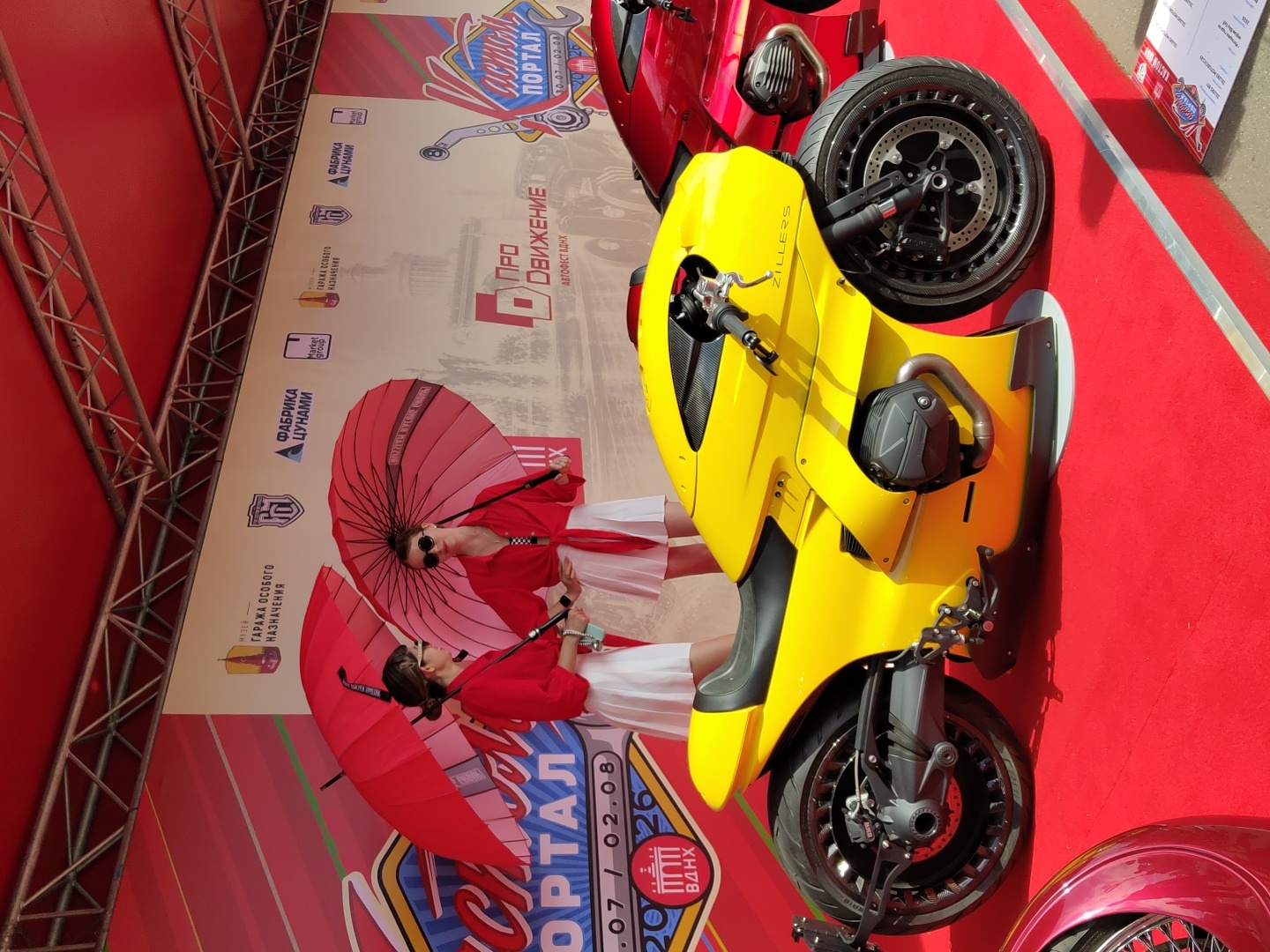 On a red carpeted stage under a red canopy, two young women stand holding large red umbrellas. They are dressed in white skirts and red blouses, with sunglasses. Behind them, a bright yellow motorcycle is parked. To the right of the yellow motorcycle, a red motorcycle is partially visible. In the background, a banner displays text in Cyrillic, including the words "ПРО ДВИЖЕНИЕ" and "Автофест". There are logos and text that read "МУЗЕЙ", "ГАРАЖА ОСОБОГО НАЗНАЧЕНИЯ", and "ФАБРИКА ЦУНАМИ", suggesting the event is at the Museum of Special Purpose Garage and a factory named TsUNAMI. The banner also includes "ВДНХ", indicating the location is VDNKh (Exhibition of Achievements of National Economy) in Moscow, Russia. The date "07 / 02.08" is also visible on the banner.