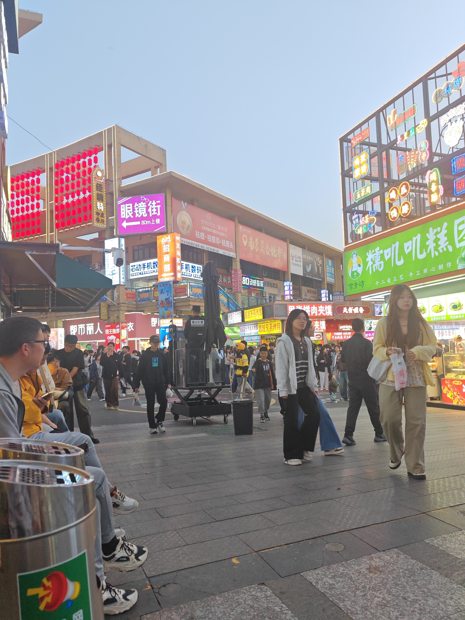 The image shows a bustling street scene in Jiangning District, China, characterized by a mix of pedestrians, shops, and signage. The street is paved with interlocking square stones. There are numerous people, including women in the foreground walking towards the camera. Many are wearing casual clothing. A man sits in the lower left corner, wearing glasses and looking at a phone. There are metal trash cans in front of him.
Buildings are lined with illuminated signs, including one that reads "眼镜街" (Yǎnjìng Jiē) indicating an eyeglasses street, and "糯叽叽糕" (Nuò jī jī gāo) for a bakery, plus a range of other commercial signs in Chinese. The signs feature neon lights and bright colors. Many of them are promoting food, and also phone service. The sky is a pale blue. Some people are walking, some are standing.