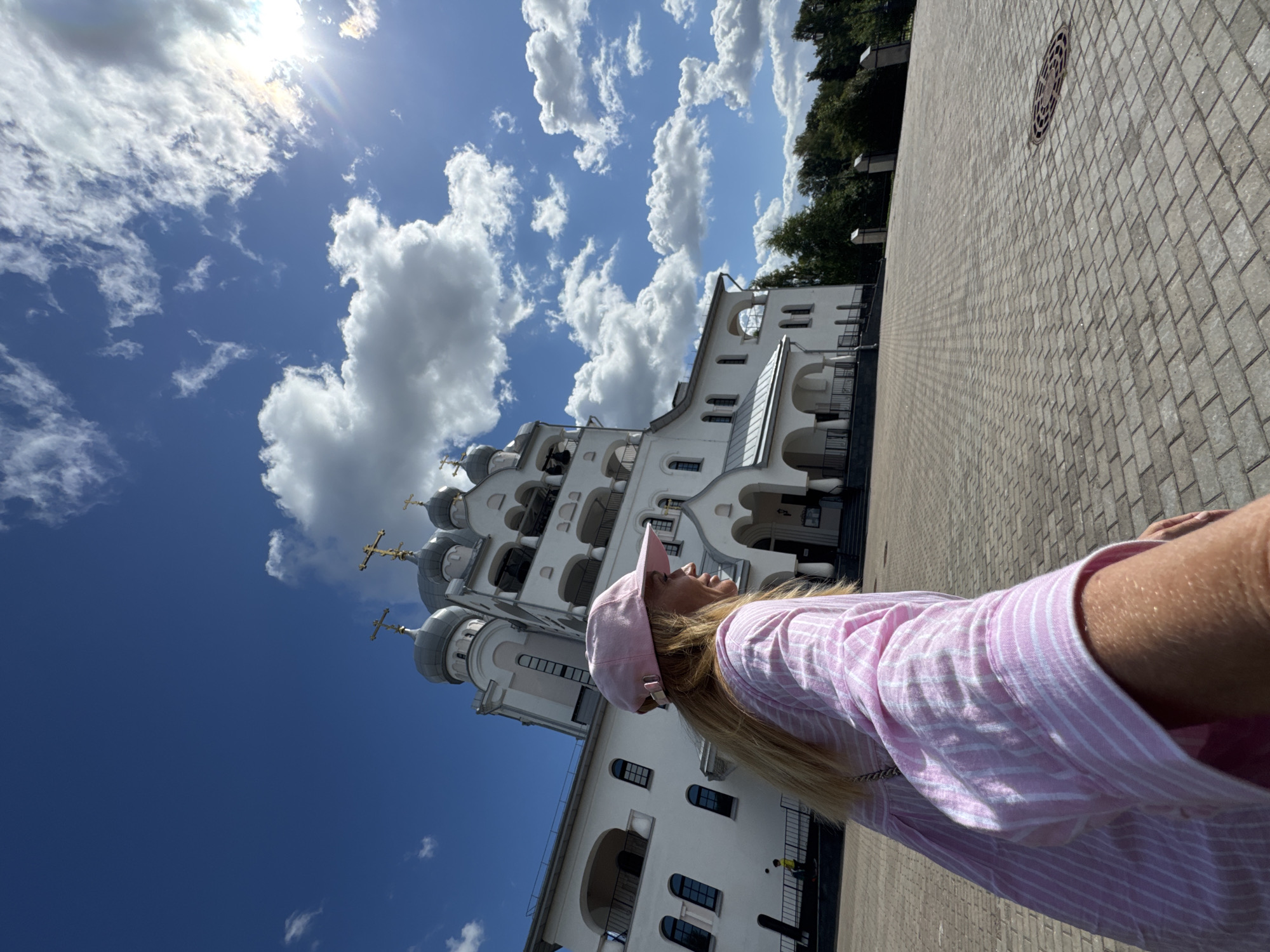 A blonde woman wearing a pink and white striped shirt and a pink baseball cap is standing in front of a white church with silver domes. The church has multiple levels and arched doorways. The woman is facing to the right, looking away from the camera. The ground in front of the church is paved with gray bricks. The sky is blue with scattered white clouds and the sun is shining brightly. The scene appears to be outdoors in an urban or suburban area, potentially Saint Petersburg, Russia, given the architectural style of the church.