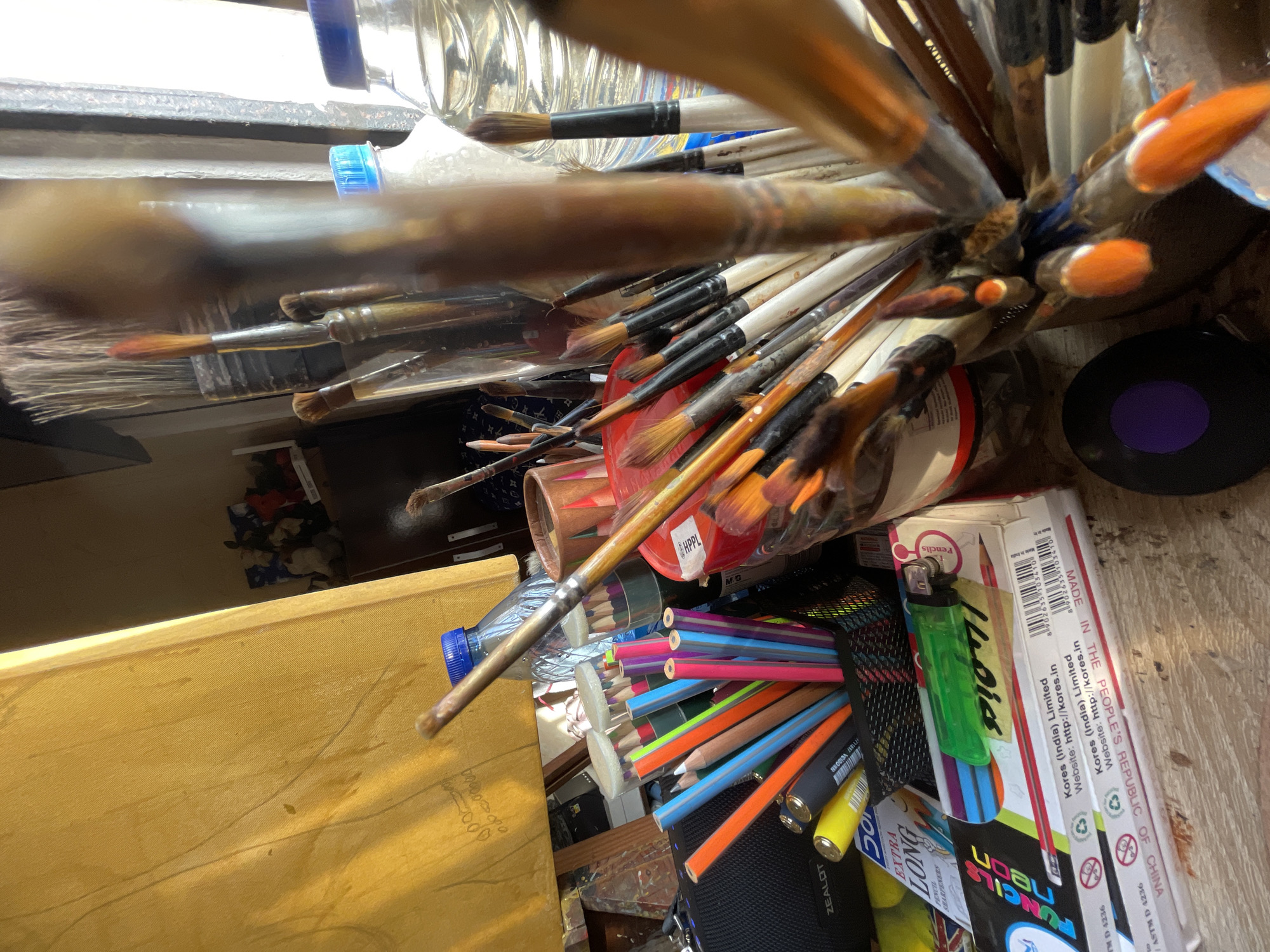 This image shows an elevated, close-up view of art supplies, including numerous paintbrushes, colored pencils, a lighter, and stationery items, crammed together in containers. The items appear to be arranged haphazardly on a surface with a wooden texture, possibly a desk or workbench. Several brands and labels, including "Kores (India) Limited," "ZEALOT," "HPPL," and "MADE IN THE PEOPLE'S REPUBLIC OF CHINA," are visible on the stationery items. Given the presence of art supplies and stationery items, the location may be an artist's studio, a classroom, or a workspace. The specific location in Ado, Nigeria, cannot be determined from the image.