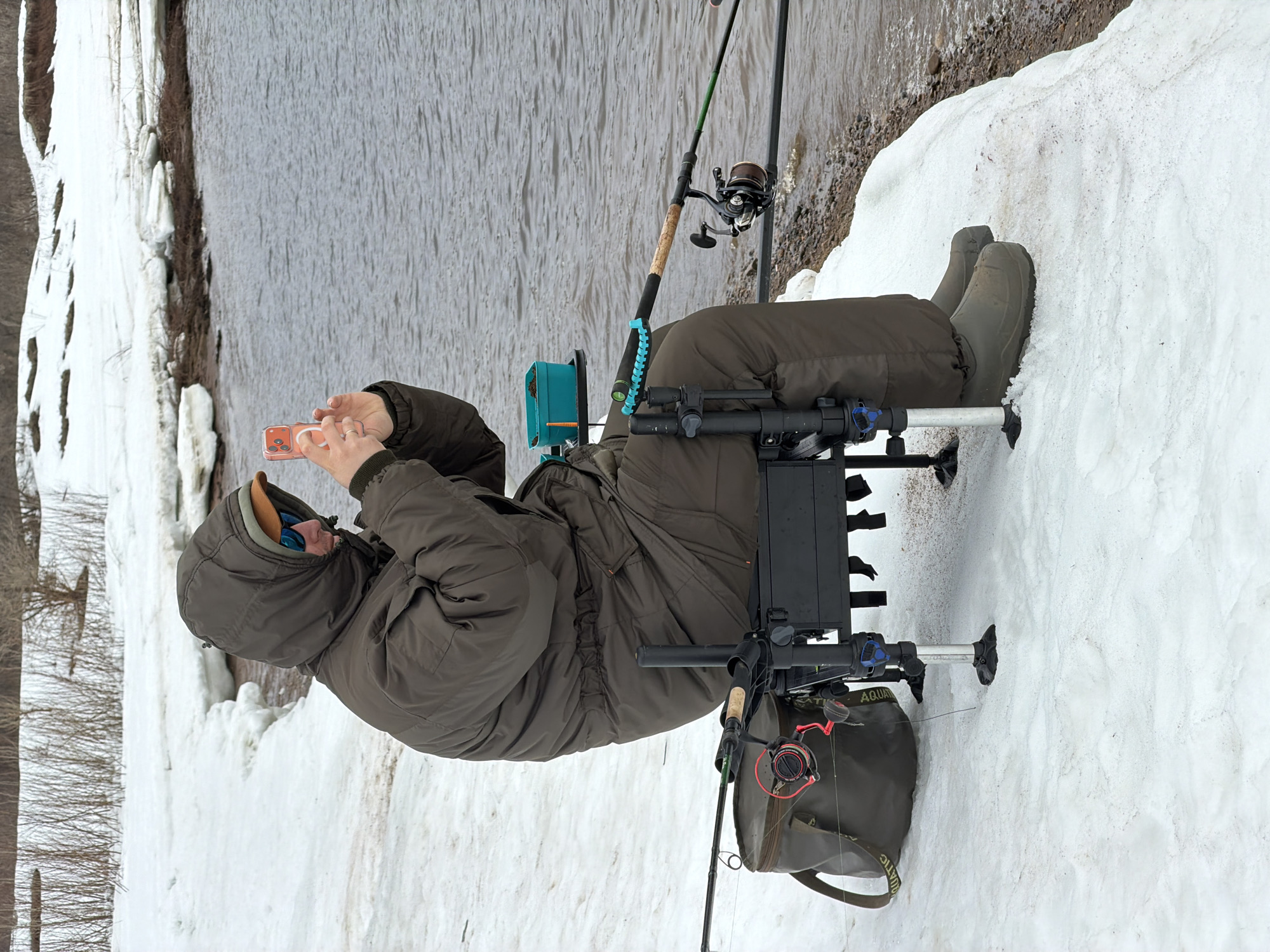 A man sits in a specialized fishing chair on a snowy bank along a river, likely in Noviy, Russia. He wears a hooded olive-green jacket, sunglasses, and matching waders. He's holding a smartphone in his hands, appearing to be taking a photo or recording a video. The fishing chair is equipped with fishing rod rests and a tackle box, suggesting a serious fishing endeavor. Two fishing rods are set up nearby. The river is visible in the background. The scene is dominated by snow and water, indicating a cold or late-winter setting.