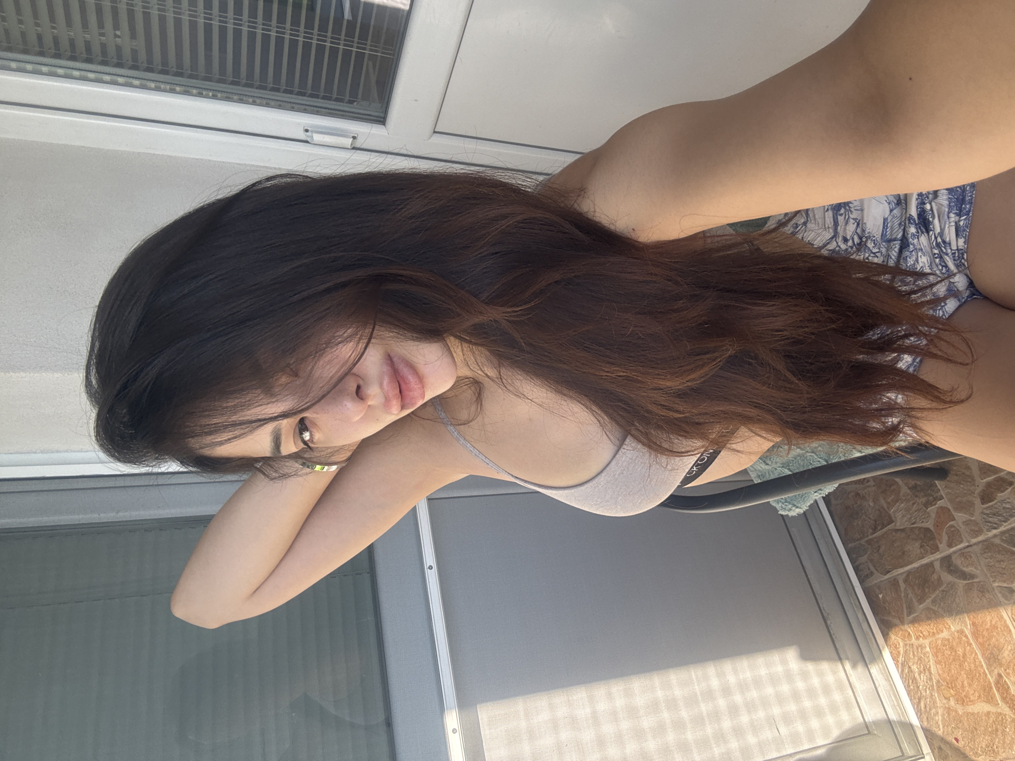 The image shows a woman sitting on a chair in an outdoor setting, possibly a balcony or patio. She has long, dark hair and is wearing a light-colored bra and patterned shorts. Her left arm is raised behind her head, and she is looking towards the camera. The background includes a window with blinds and a tiled floor. There is no specific information in the image to suggest the location is Plovdiv, Bulgaria.