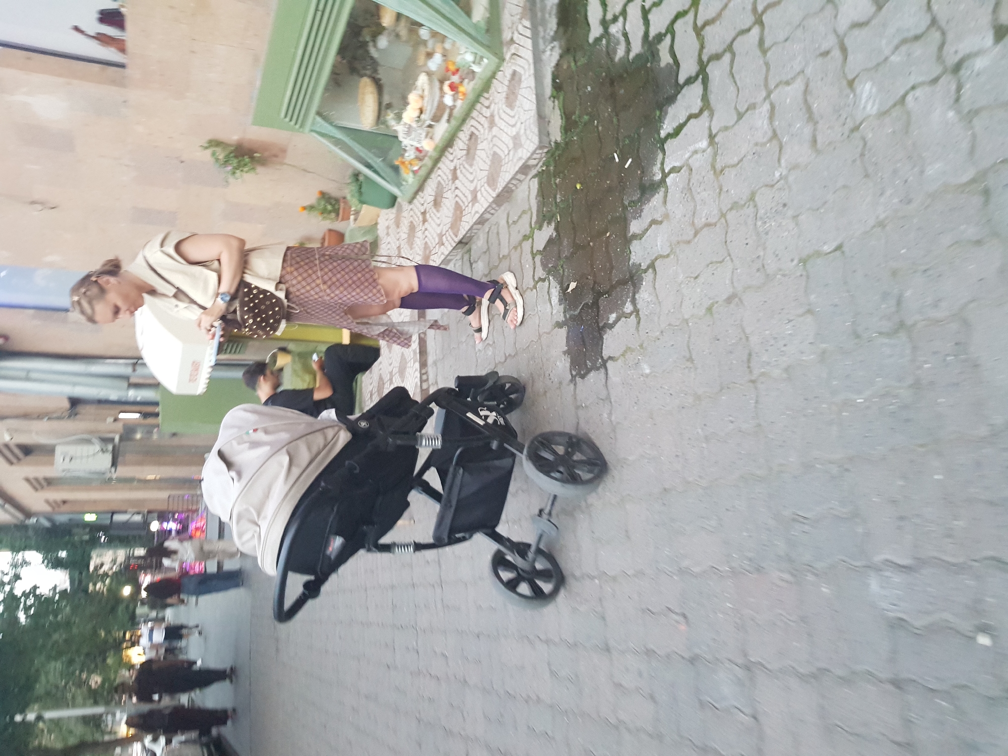 In an outdoor urban setting, a woman stands near a parked stroller on a paved sidewalk. She is wearing a light-colored sleeveless vest over a patterned skirt and dark purple knee-high socks, with sandals on her feet. A crossbody bag with gold studs is slung across her body. She appears to be looking down at a white object in her hands. The stroller is beige and black, positioned in the foreground to the left of the woman. In the background, several other people are visible walking on the sidewalk, and a building with a textured wall stands behind them. There is a green display case or window with plants and decorative items in front of the building. The ground is a mosaic of interlocking grey pavers, with some wet patches and debris.