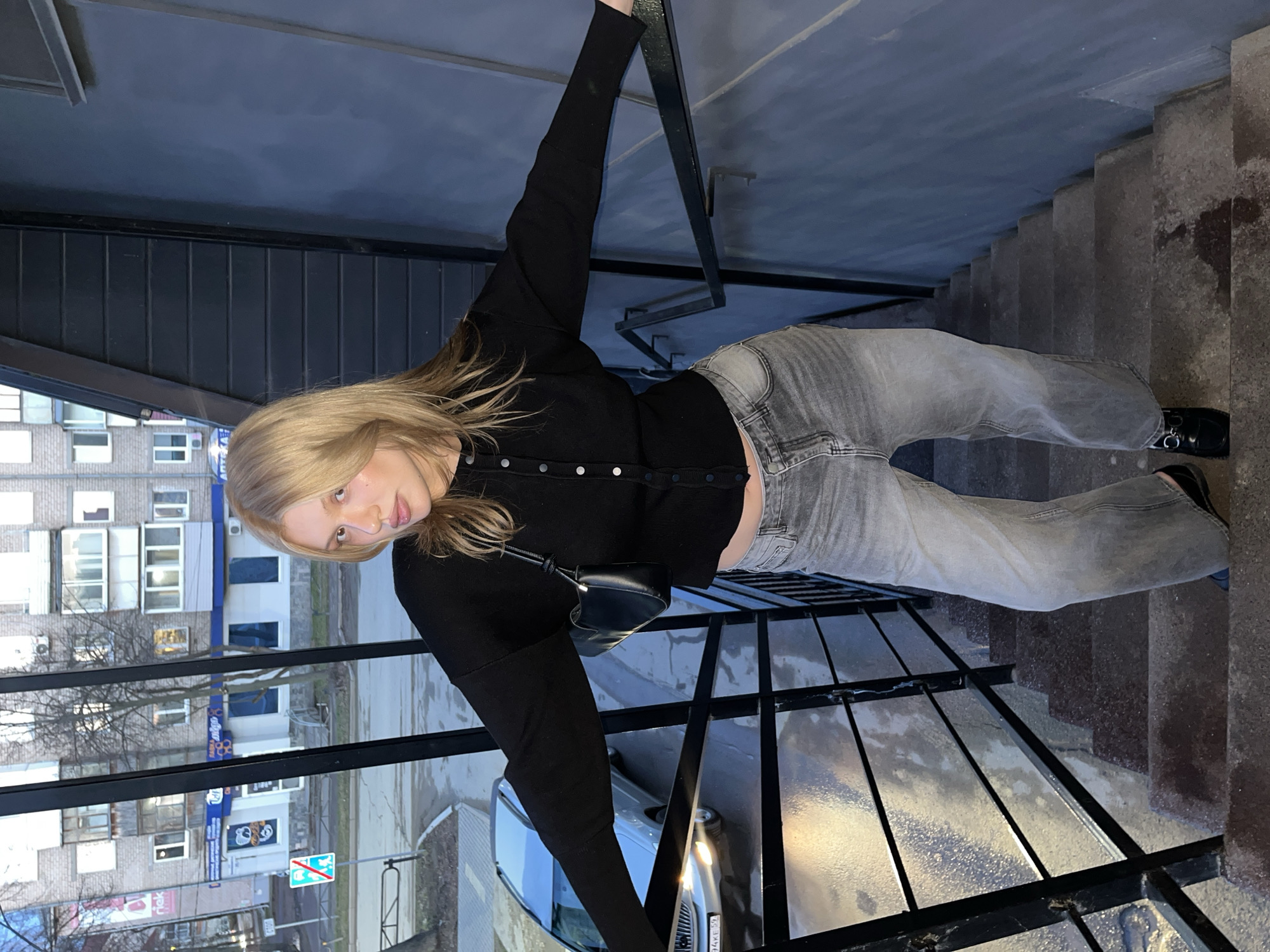 A young woman with long blonde hair, dressed in a black long-sleeved button-up top and light grey jeans, poses in an urban setting, possibly an exterior stairwell or balcony in Chaikovsky, Russia. She is shown in a dynamic pose, holding onto a black metal railing with her right arm extended upwards, looking directly at the camera with a neutral expression. A small black shoulder bag is slung across her body.
The setting features concrete stairs descending to the right, and sturdy black metal railings forming a barrier. Surfaces appear wet, suggesting recent rain or damp conditions. In the background, beyond the railings, several multi-story residential buildings with various windows and balconies are visible. Leafless trees line what appears to be a street or path. A parked car with illuminated headlights is partially visible below the woman.
The ambient lighting suggests dusk or evening, with some artificial lights visible on the buildings and from the car. A blue sign in the background features white text that includes "Аптека" (Pharmacy) and "Открыто 24 часа" (Open 24 hours). Another road sign, possibly indicating a parking or stopping restriction, is also present. The overall scene depicts a casual moment within a somewhat utilitarian outdoor urban space.