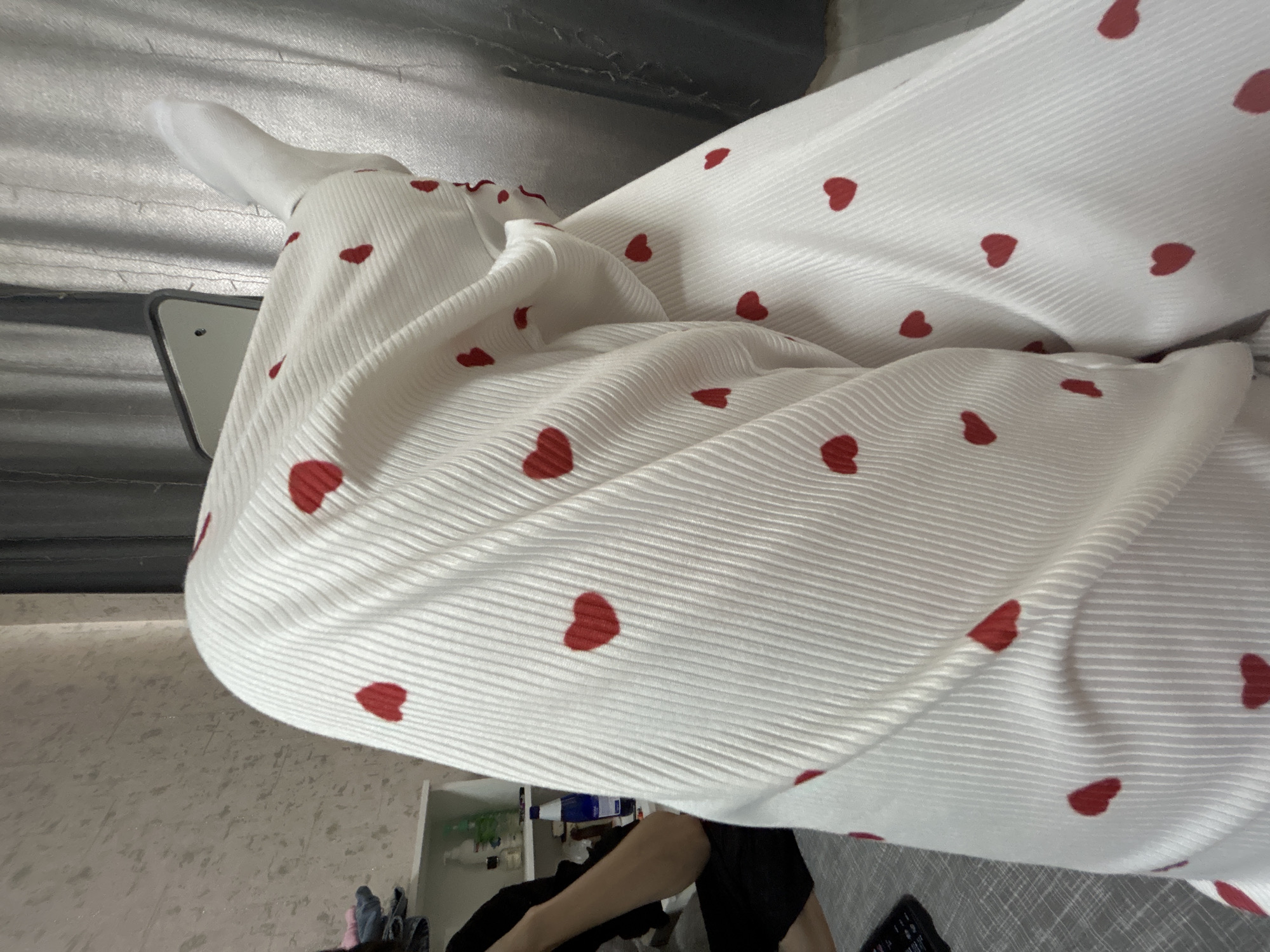 A white, pleated fabric with red heart patterns is prominently featured. A white sock is visible at the top left. The fabric partially obscures a view of a person wearing black clothing. A shelving unit with various items on it is visible in the background, along with a wall and a silver-colored ceiling. The location and its city cannot be determined from the image.