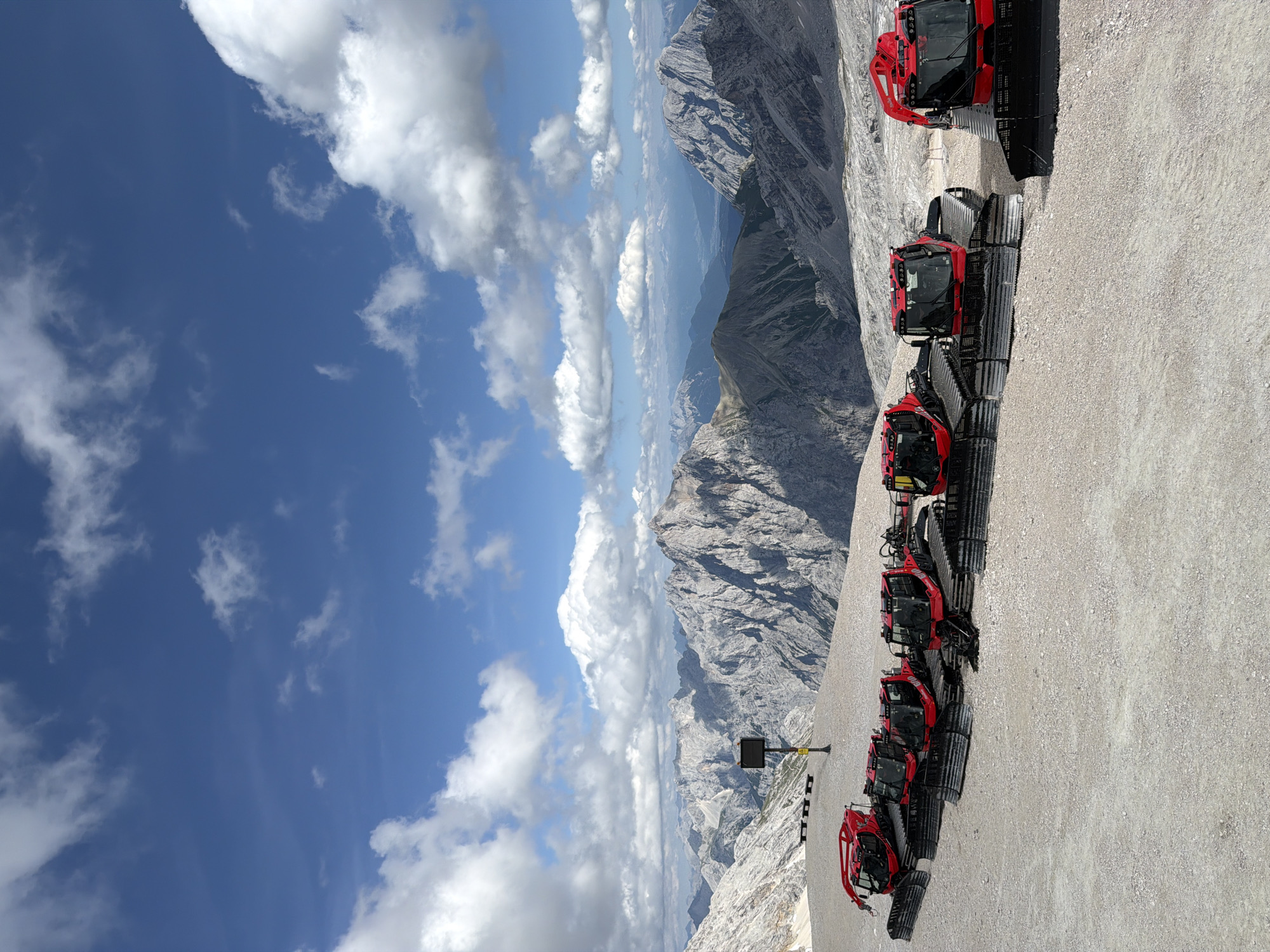 In the city of Garmisch-Partenkirchen, Germany, on a sunny day with scattered clouds, a line of red snowcats is parked on a gravelly, mountainous terrain. The snowcats are positioned diagonally across the frame, facing to the left. Behind them, a dramatic mountain range stretches into the distance under a vibrant blue sky. The mountains are rugged and rocky, with some appearing snow-capped. A black sign on a pole stands to the left of the snowcats, and further back, a few wooden benches are visible. The overall scene depicts a high-altitude, possibly tourist-oriented, location in the Bavarian Alps.
