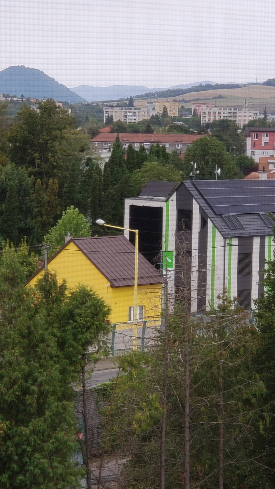 The image shows a view of a residential area in the city of Prešov, Slovakia. In the foreground, there is a bright yellow house with a brown roof and a single visible window. Adjacent to the yellow house, a modern building with a black and white striped facade and solar panels on its roof is visible. The building features vertical green stripes. Further in the background, several multi-story apartment buildings and other structures can be seen, nestled amongst trees. The distant landscape is characterized by rolling hills and mountains under a cloudy sky. The scene depicts a mixed urban and natural environment.