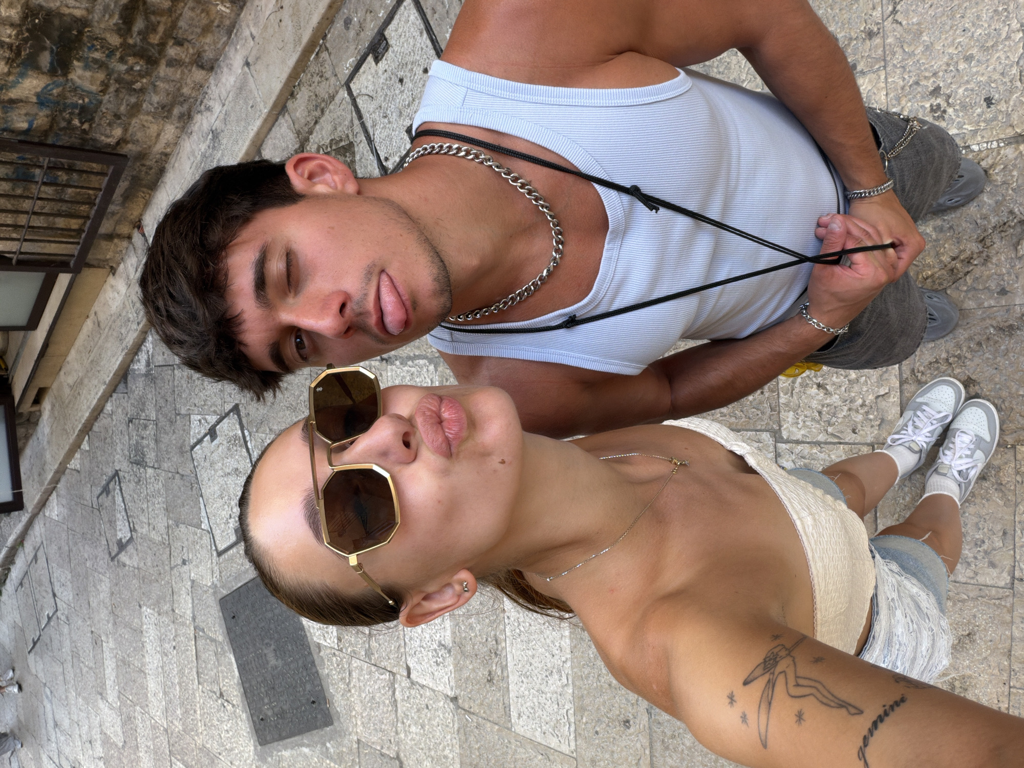 This is a high-angle, full-body selfie taken by two young adults in what appears to be a cobblestone alleyway in Bari, Italy. The woman on the left is wearing a white off-the-shoulder top, denim shorts, and white sneakers. She has on large, gold-rimmed sunglasses and her lips are pursed in a kissing gesture. A delicate silver necklace adorns her neck. On her left arm, a tattoo of a fairy and the word "gemini" are visible. The man on the right is wearing a light blue ribbed tank top, grey pants, and grey slides. He is winking and sticking his tongue out. A thick silver chain necklace is around his neck, and a black cord is also visible. He has a silver bracelet on his left wrist and another chain attached to his pants. The background shows a textured stone wall and metal bars, consistent with historic Italian architecture. The ground is paved with irregular grey stones, and a couple of manhole covers are visible. The lighting suggests it is daytime.