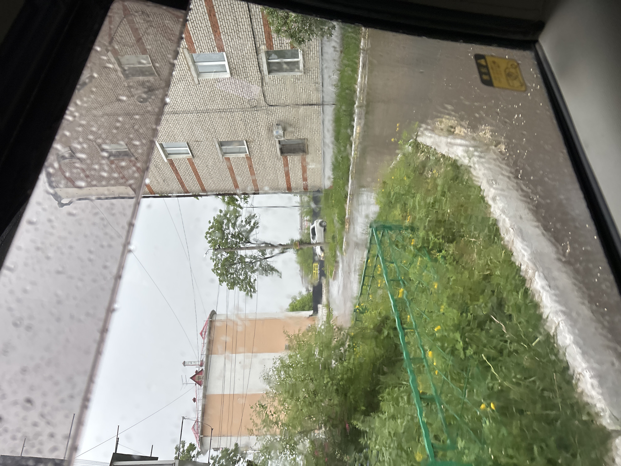 The image shows a view through a rain-streaked window. Outside, there's a multi-story brick building with white-framed windows and some red brick accents. A sidewalk runs alongside this building. Further away, there's a taller, pink building with white stripes and a red roof, partially obscured by trees and bushes. A single tree stands between the buildings, and a white car is parked on the wet street. A green fence is visible in the foreground among the vegetation.