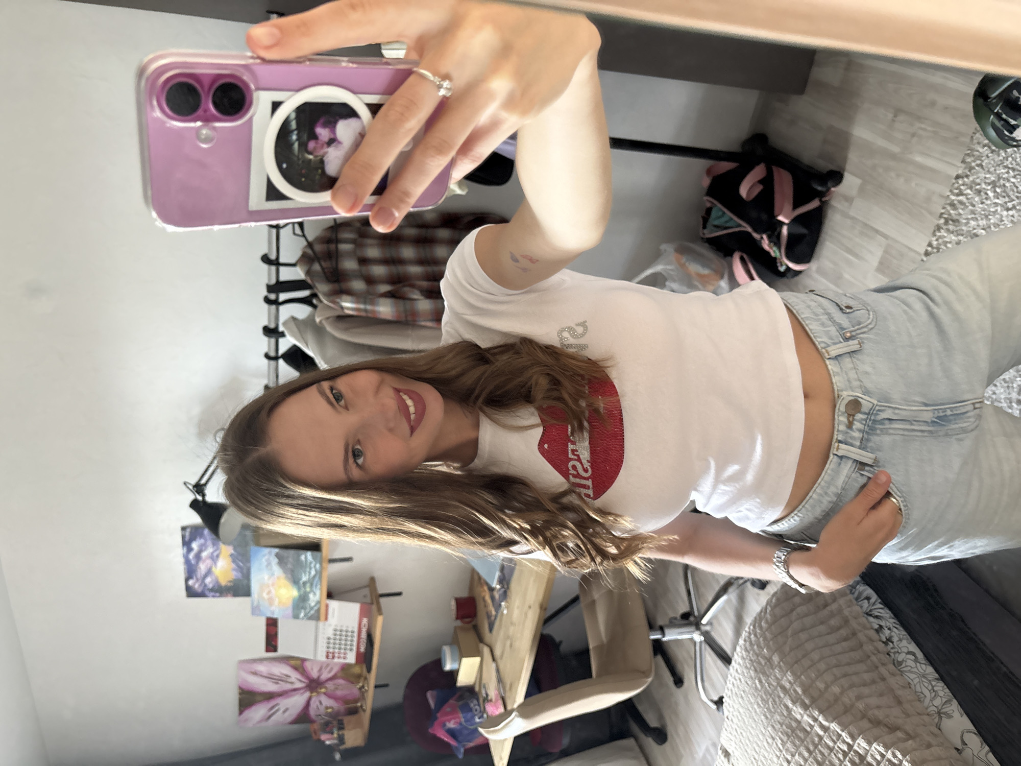 A young woman is taking a mirror selfie in what appears to be her bedroom. She is wearing a white t-shirt with a red sequined lip design on it and light-wash denim jeans. The woman has long, wavy brown hair and is smiling at the camera. She is wearing a silver ring on her left hand and a silver wristwatch on her right wrist.
In the background, the room is somewhat cluttered. There is a desk with art supplies, paintings, and a calendar on it. A black desk lamp is attached to a shelf above the desk. A black backpack with pink accents is visible on the floor. A plaid shirt is hanging on a rack. The overall impression is of a lived-in, personal space, possibly reflecting the woman's artistic interests. The lighting suggests it is daytime, with natural light coming from an unseen window. The location context provided is Perm, Russia.
