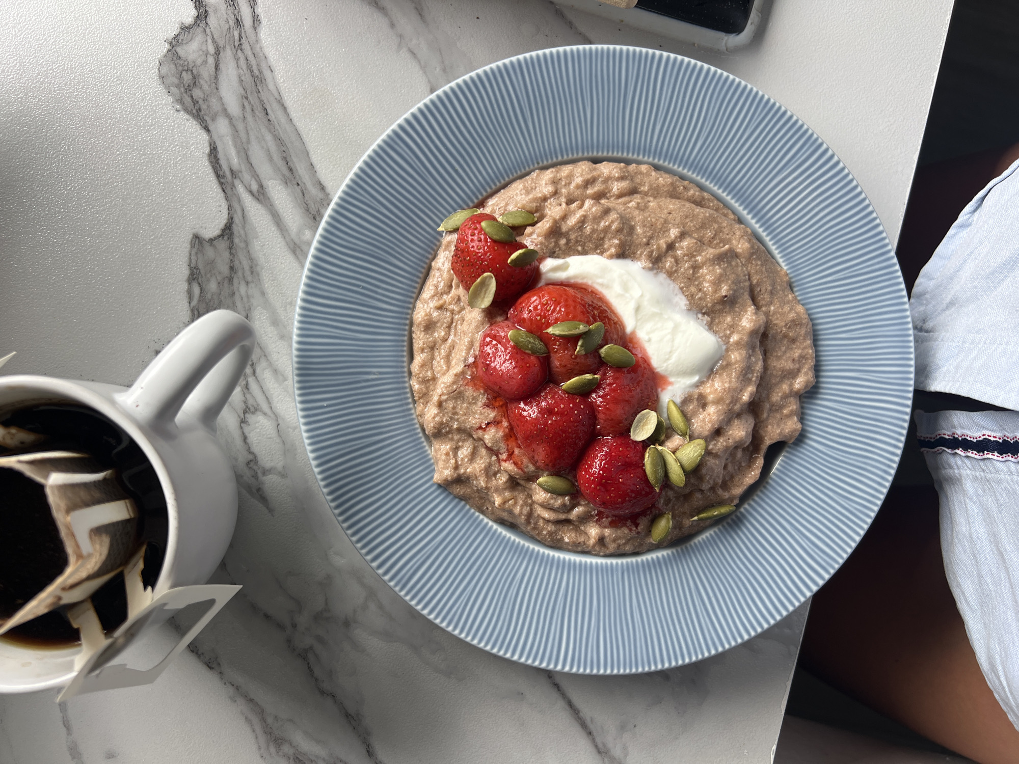 A close-up overhead shot shows a light blue plate with a ribbed pattern filled with oatmeal topped with strawberries, yogurt, and pumpkin seeds. Next to the plate is a white mug filled with dark coffee and a drip coffee filter. The objects are placed on a white marble-patterned surface. The lighting suggests a bright, natural light source coming from the top left. The composition is focused on the food and drink, with a person's legs and clothing visible at the bottom of the frame.