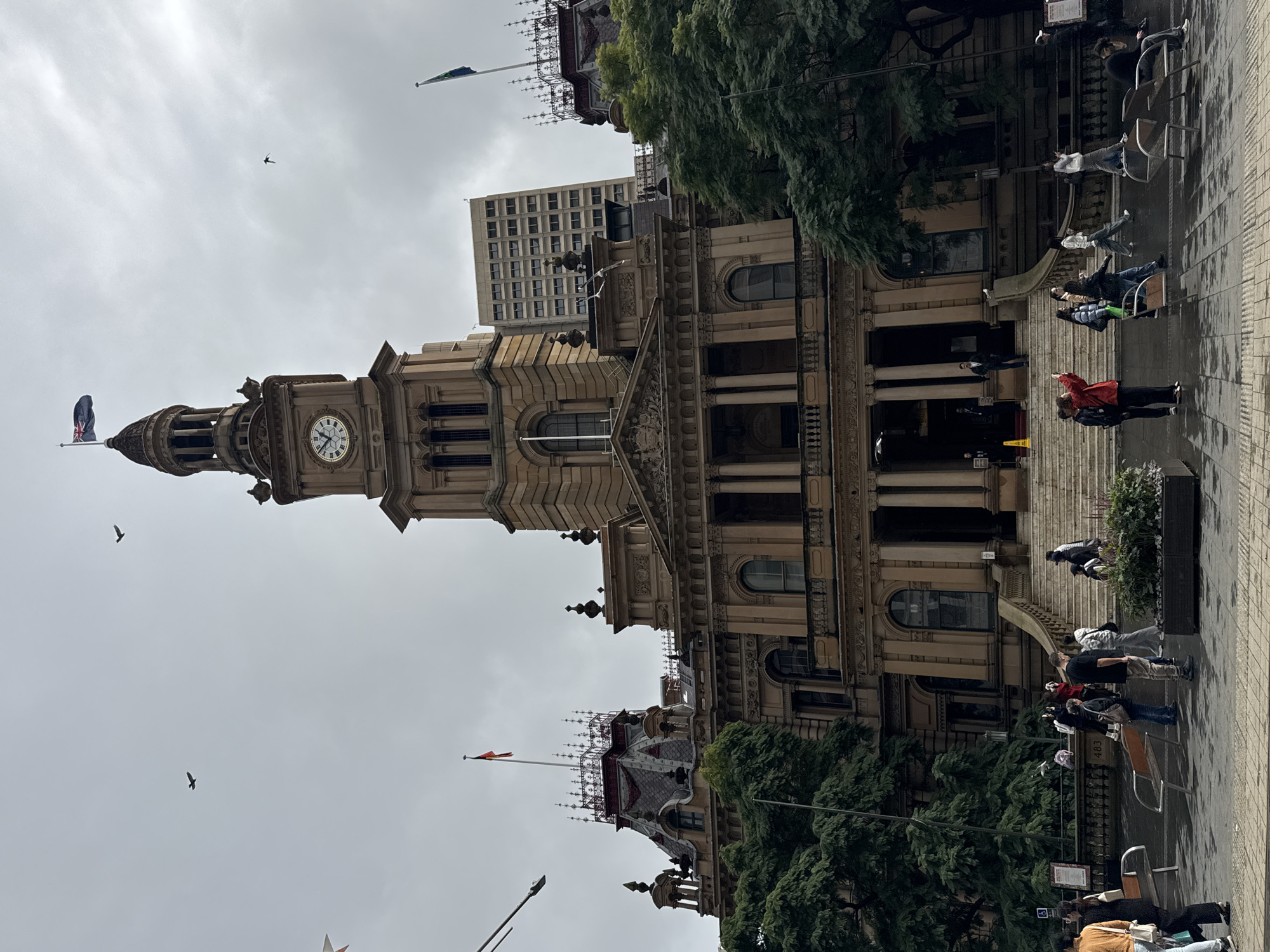 The image captures a frontal view of a large, ornate building with a prominent clock tower, likely a historic civic structure, under an overcast sky. Several birds are visible in flight against the sky. In the foreground, a paved plaza is wet, suggesting recent rain. A number of people are present in the plaza, some walking, some standing, and a few appear to be taking photographs. The building features classical architectural elements, including columns, arched windows, and decorative stonework. Trees with dense green foliage flank the building. The overall atmosphere is muted due to the cloudy weather.