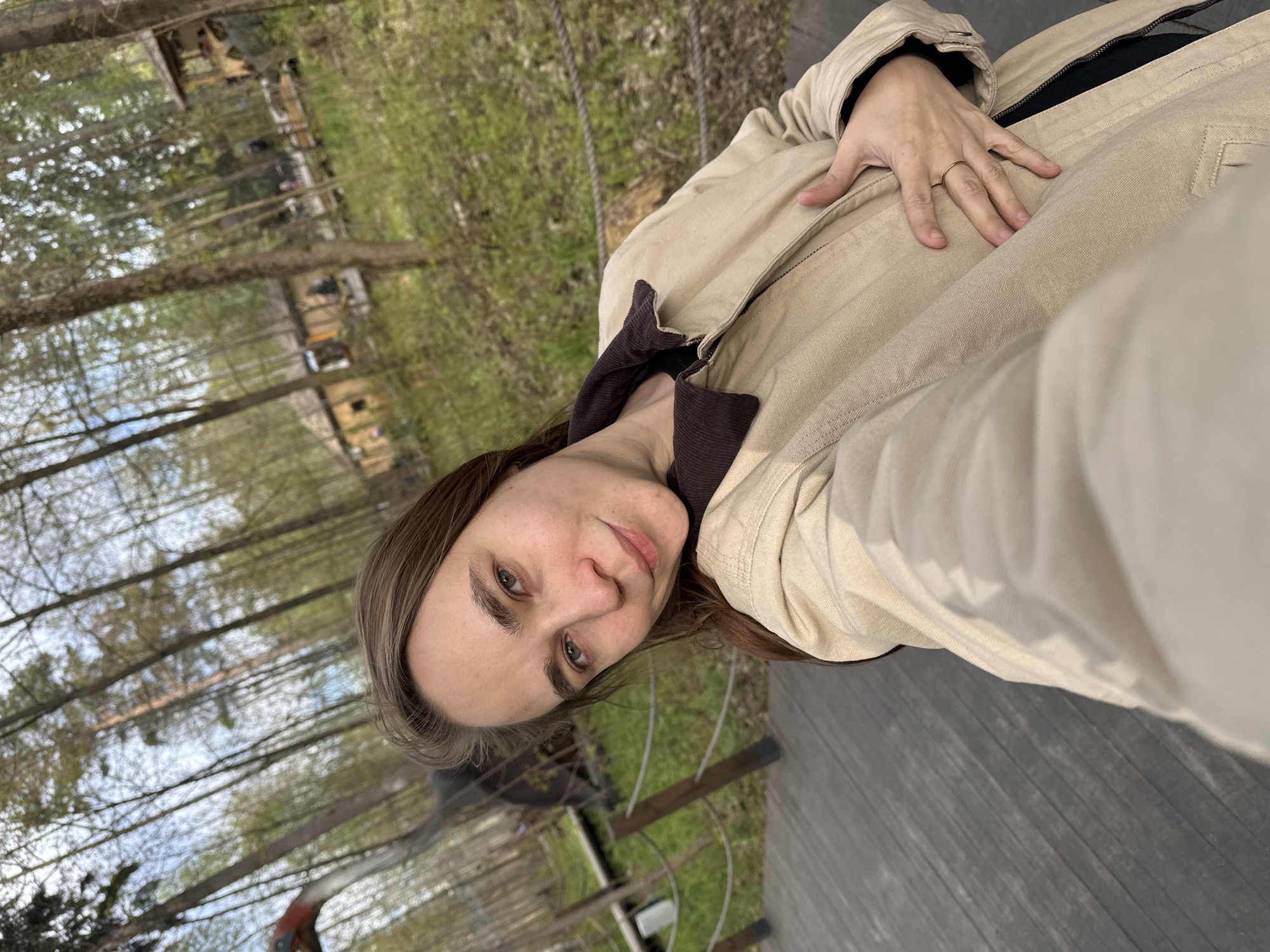 A woman is taking a selfie. She is wearing a beige jacket over a dark top. Her hair is light brown. She has light skin and green eyes. She is wearing a ring on her left ring finger. The background shows a wooded area with tall trees. There is a wooden walkway in the foreground. The location is Izhevsk, Russia.
