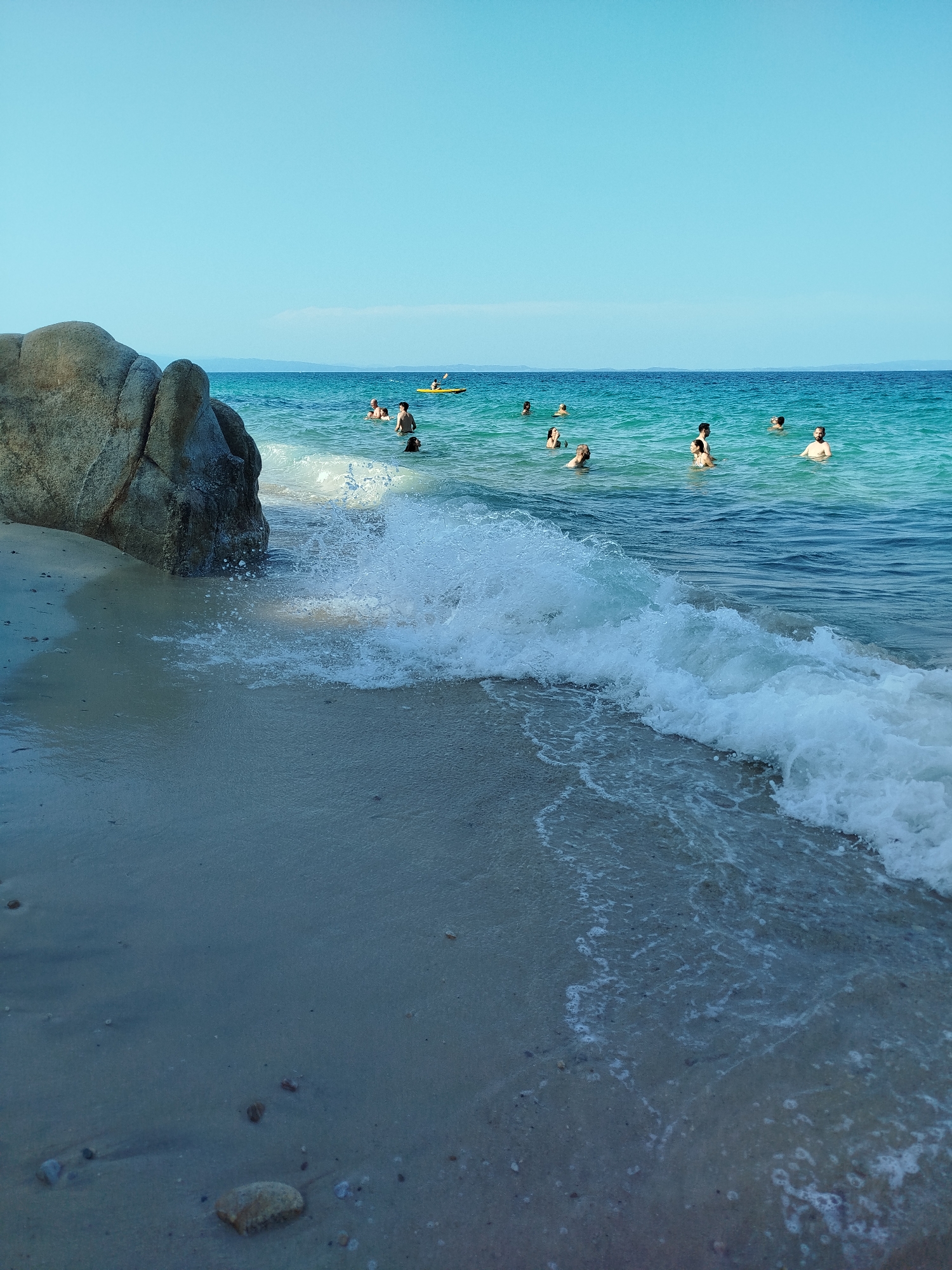 This is a scene of a beach in the Sithonia Municipal Unit, Greece. In the foreground, a wave is breaking on the sandy shore, with foam and small pebbles visible on the wet sand. To the left, a large, weathered rock formation juts out towards the water. In the middle ground, the turquoise sea is populated by numerous swimmers. One person is visible in a yellow kayak in the distance. The sky is a clear, bright blue. The overall impression is a sunny day at a popular Greek beach.
