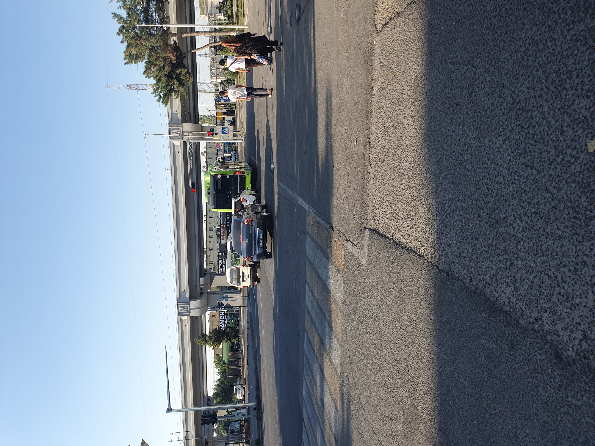 This is a street-level, eye-level shot of an intersection in Tashkent, Uzbekistan, on a clear, sunny day. A pedestrian crossing is visible in the foreground. In the middle ground, a green bus and a dark sedan are stopped at a traffic light, with a white van also visible. Three pedestrians are walking away from the camera across the intersection, and another pedestrian is walking towards the camera. An elevated roadway passes overhead, supported by concrete pillars. In the background, several buildings and trees are visible. The overall impression is of a busy urban intersection.