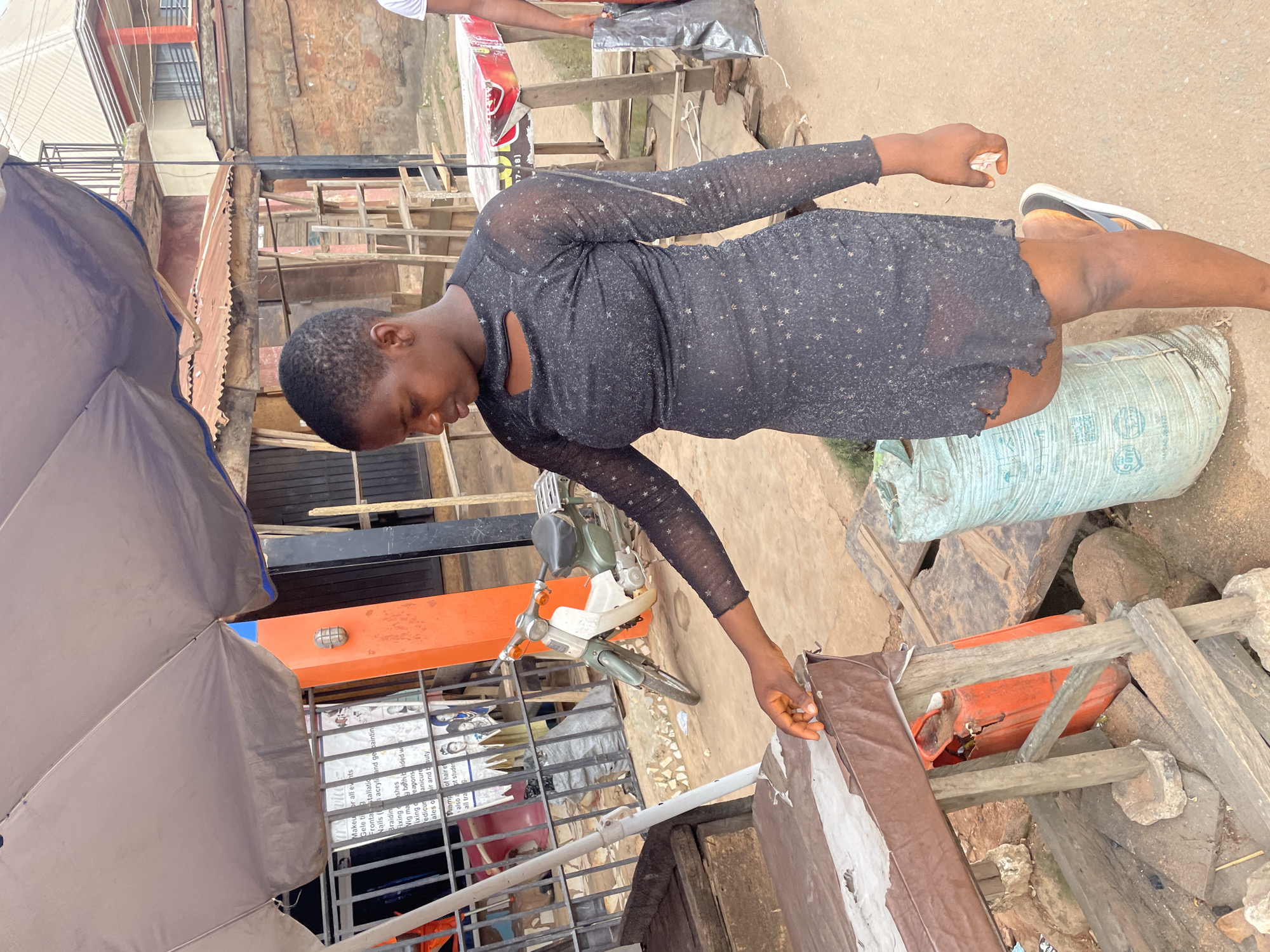 A young woman, appearing to be in her late teens or early twenties, stands outdoors in Nkpor, Nigeria, near a market stall. She is wearing a sheer, black, long-sleeved dress with a star pattern. Her hair is short and dark, and she is looking down towards a brown table with a torn, light-colored surface. Her right hand is extended, touching the edge of the table. Beside her, on the ground, is a large, light blue, woven sack. In the background, a green and white motorcycle is parked, and a building with an orange-colored pillar is visible. Other market stalls and structures are present in the immediate vicinity. The ground is paved with dirt and small stones.