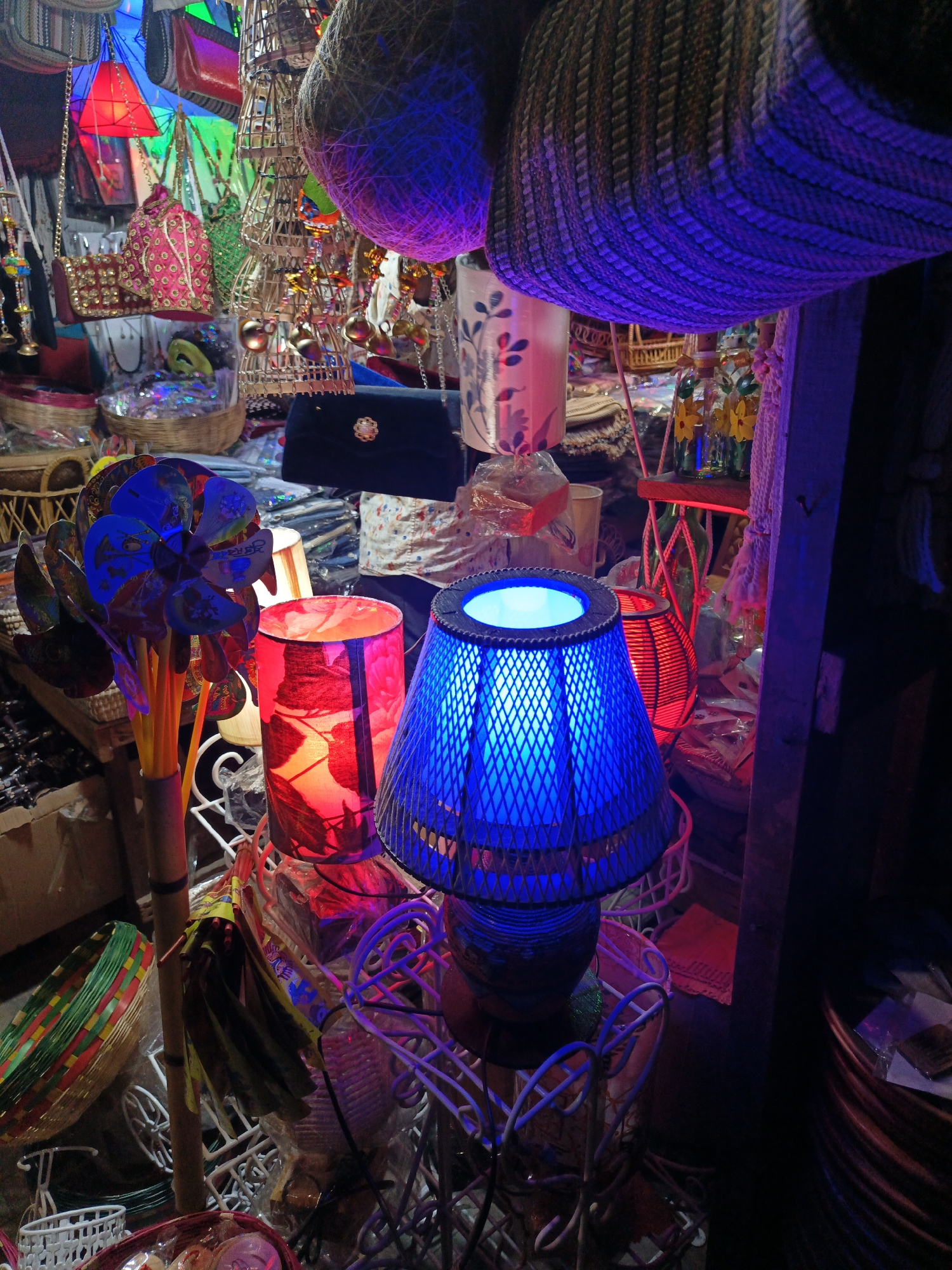 This image, likely taken in Dhaka, Bangladesh, shows a cluttered indoor marketplace. The foreground features a blue, lattice-shade table lamp, positioned next to a red cylindrical lamp. A bunch of blue pinwheels on sticks stand nearby. Overhead are hanging baskets. In the background are various items for sale, including purses, bags, ornaments, and decorations. The lighting is varied with the blue and red lamps casting strong colored glows.