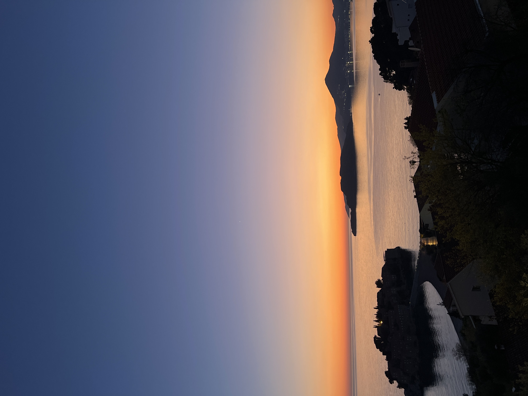 The image depicts a landscape at sunset, likely taken from the city of Sveti Stefan, Montenegro. The sky is transitioning from deep blue on the left to a vibrant orange towards the horizon on the right. A body of water reflects the colors of the sunset, creating a shimmering effect. In the distance, a dark silhouette of a mountain range is visible with scattered lights indicating possible settlements. Closer to the foreground on the right, there are structures including houses with dark rooftops and trees. These features are also silhouetted. The general environment appears calm and serene.