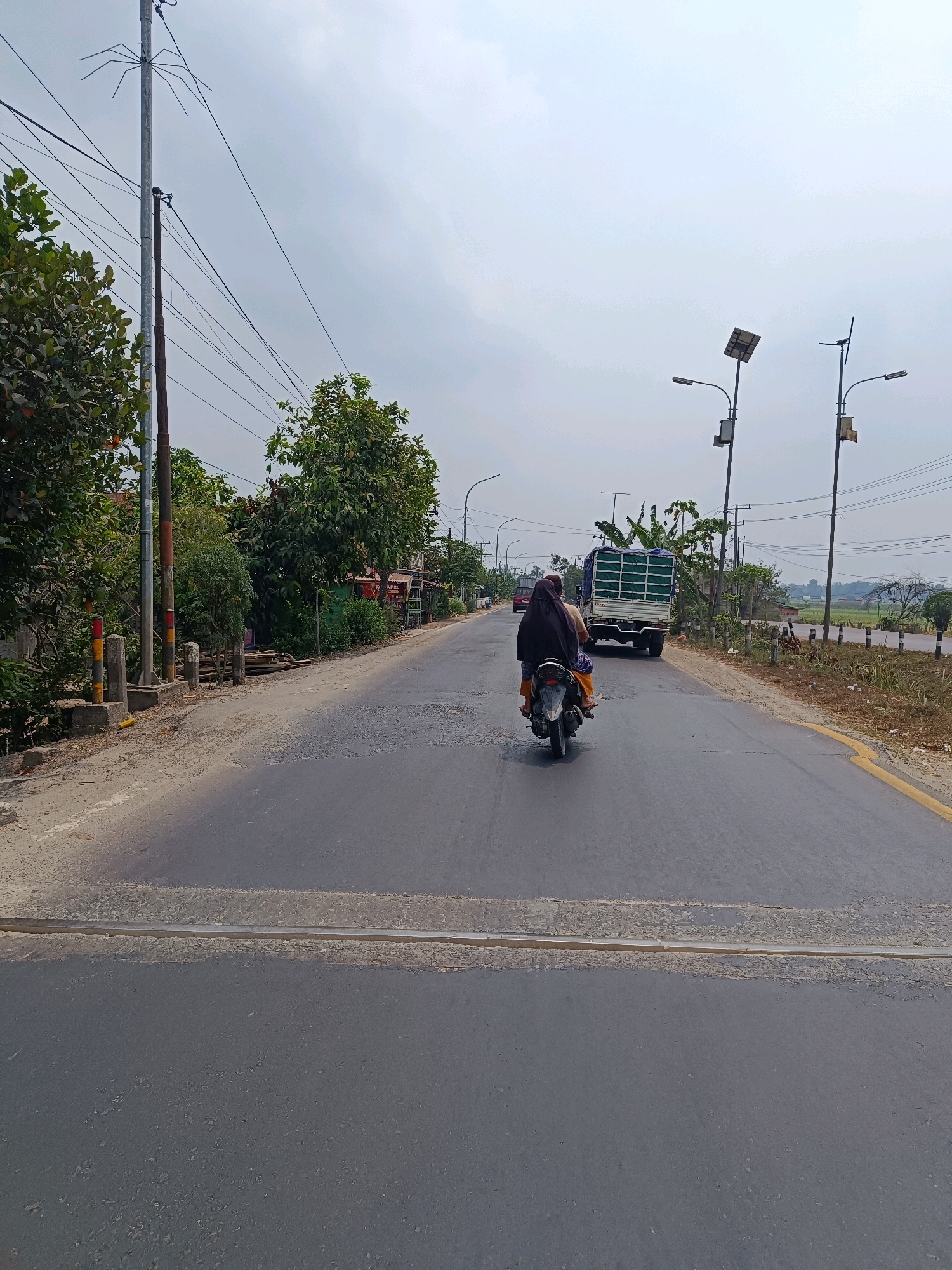 This is a photograph taken on a cloudy day, showing a road in the city of Kayu Agung, Indonesia. The road is asphalt, with a yellow line marking its edge on the right side. In the foreground, there is a pedestrian crossing marked by two light gray stripes. A woman wearing a black headscarf and orange pants is riding a motorcycle away from the camera, her body angled to the right. Behind her, a white truck with a green tarp covering its cargo is driving in the same direction.
On the left side of the road, there are trees and vegetation, with a utility pole that has a spider-like structure attached to its top. Further down the road, more trees and buildings are visible, some appearing to be shops. On the right side of the road, there is a field and more utility poles with solar panels. The sky is overcast with light gray clouds. The overall scene depicts a typical roadside environment in a moderately developed area.