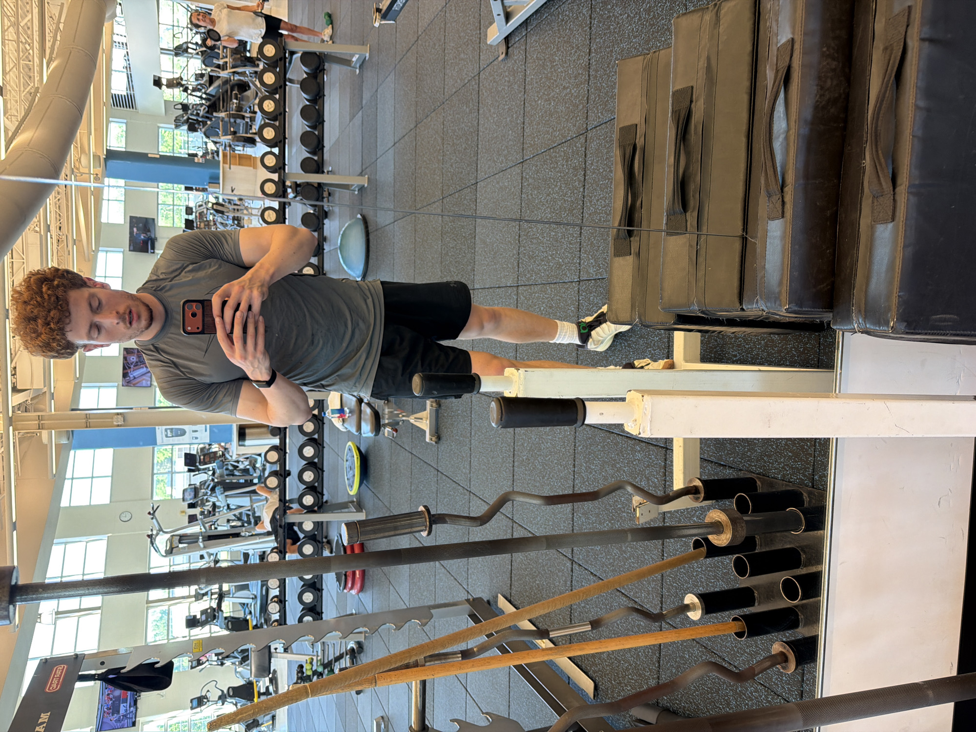 A man with curly reddish-brown hair is standing in a gym, taking a mirror selfie with his smartphone. He is wearing a dark grey t-shirt and black athletic shorts. His left arm is raised, holding the phone, while his right hand is partially visible in front of the phone. He is looking at his reflection.
The gym environment is visible in the background and through the mirror. There are various exercise machines, including treadmills and ellipticals, as well as weight racks with dumbbells and barbells. In the background, to the right of the man, a woman is visible on a treadmill. To the left of the man, in the foreground, a rack of barbells and other weightlifting equipment is present. A stack of black weight pads is in the lower right corner of the image. The floor is covered with black rubber gym flooring. The gym appears to be well-lit with natural light coming from large windows and artificial lighting. The location is identified as Beachwood, United States of America.