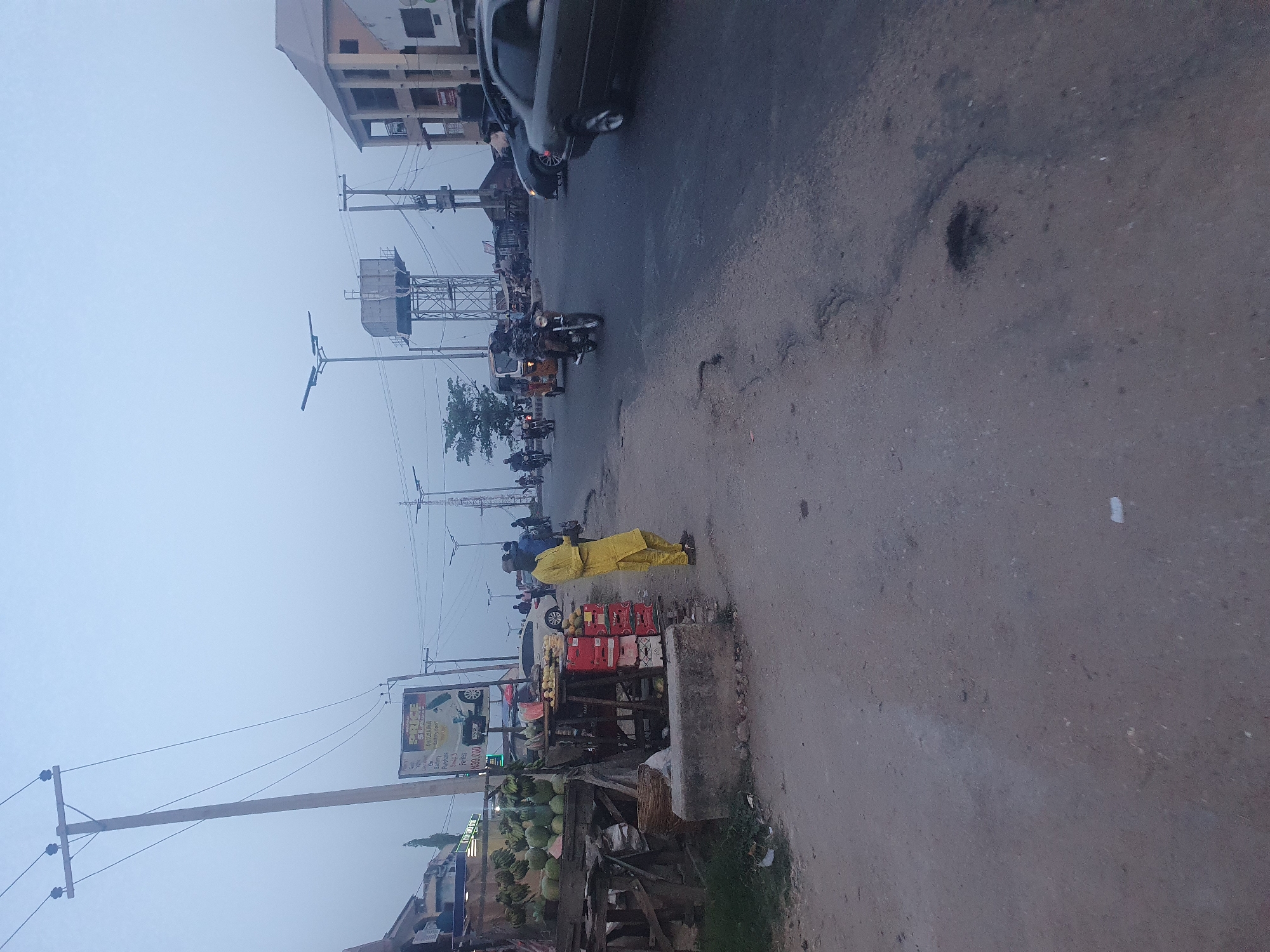 The image depicts a street scene in Lokoja, Nigeria, under overcast skies. A man in a yellow robe is walking on the shoulder of the road next to a roadside fruit stand. The stand has coconuts, bananas, oranges, and red Coca-Cola crates. A sign is beside the stand. Several motorcycles and a dark car are driving on the road in the distance. A large water tower is visible behind the vehicles. Buildings and electrical lines are present on both sides of the road. The road surface shows signs of wear and tear.