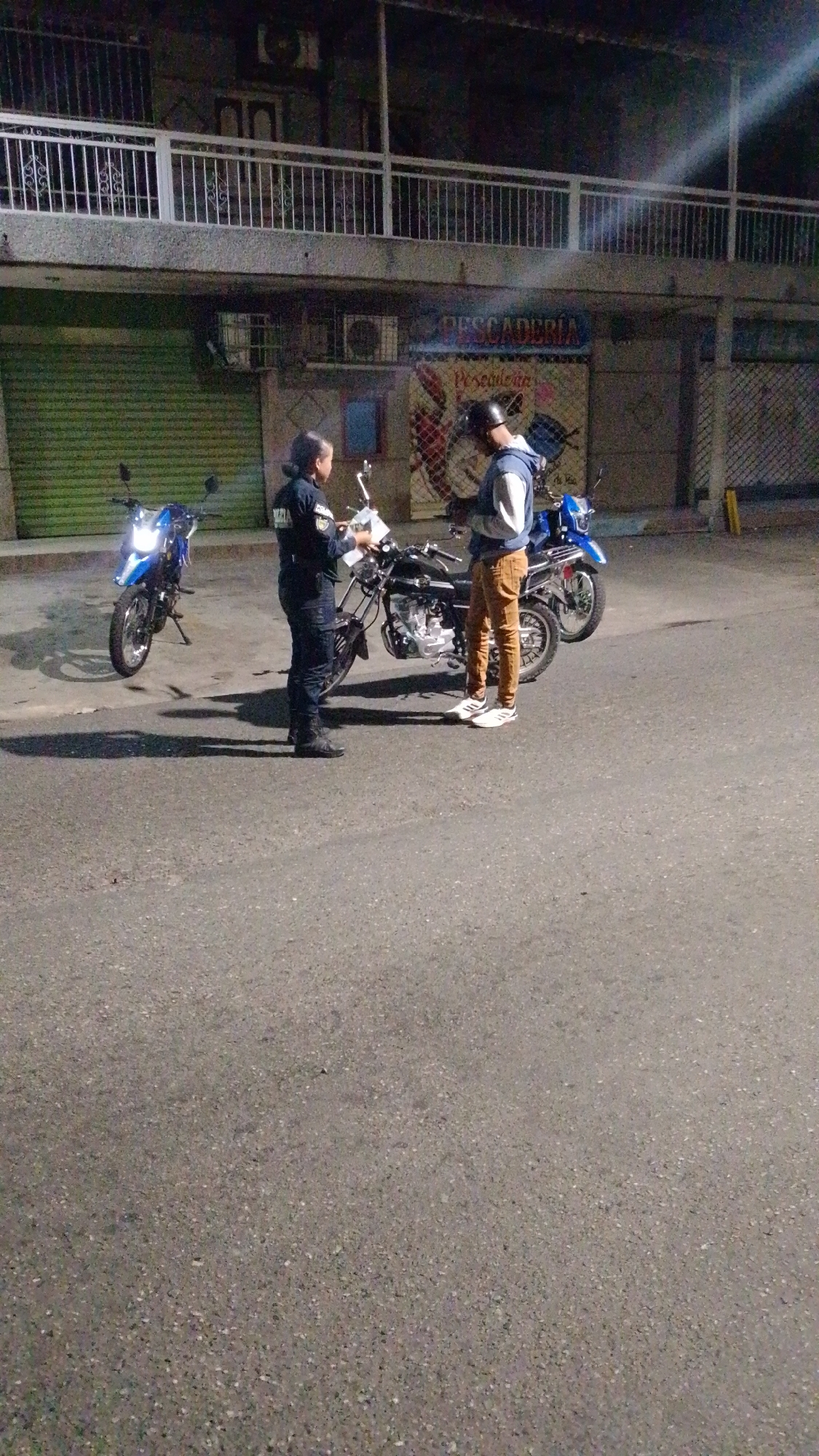 In the city of Tinaquillo, Venezuela, a female police officer in uniform is seen interacting with a male civilian wearing a blue jacket and tan pants. They stand next to a black motorcycle on a paved street. Behind them, a building with a metal balcony and closed storefronts is visible. Two additional motorcycles are parked nearby, one blue and one partially obscured. The scene appears to be at night or in low light conditions. The officer is holding papers, possibly issuing a ticket or documenting information.